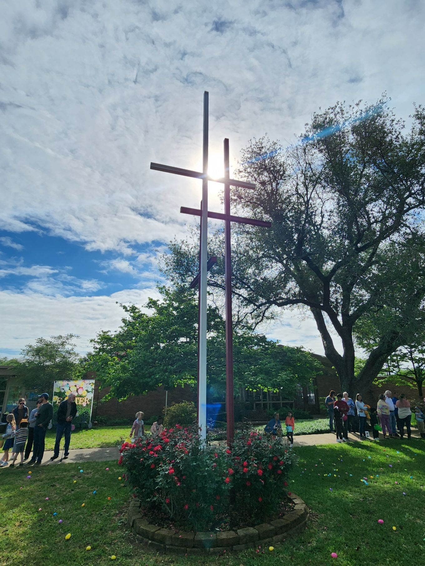 Three crosses with sun shining behind; people gather on green grass.