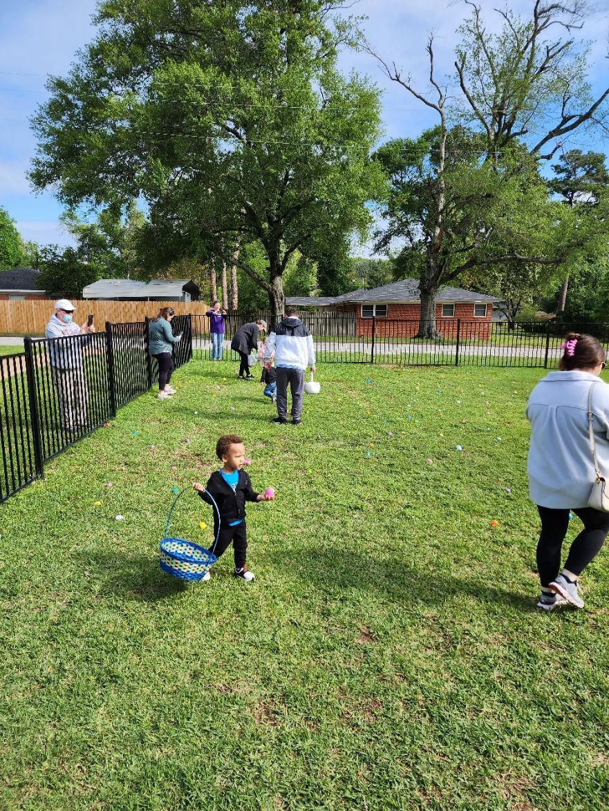 Children hunting for Easter eggs in a grassy yard, with adults watching. Black fence and houses in the background.
