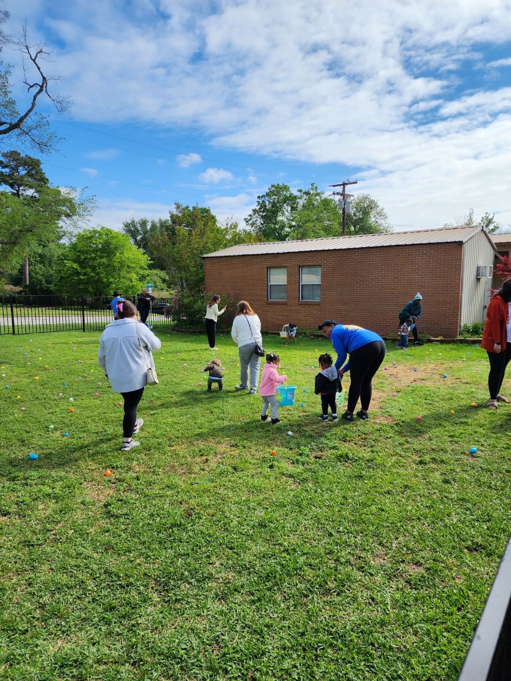 People and children hunt for Easter eggs in a grassy yard, with a building and trees in the background.