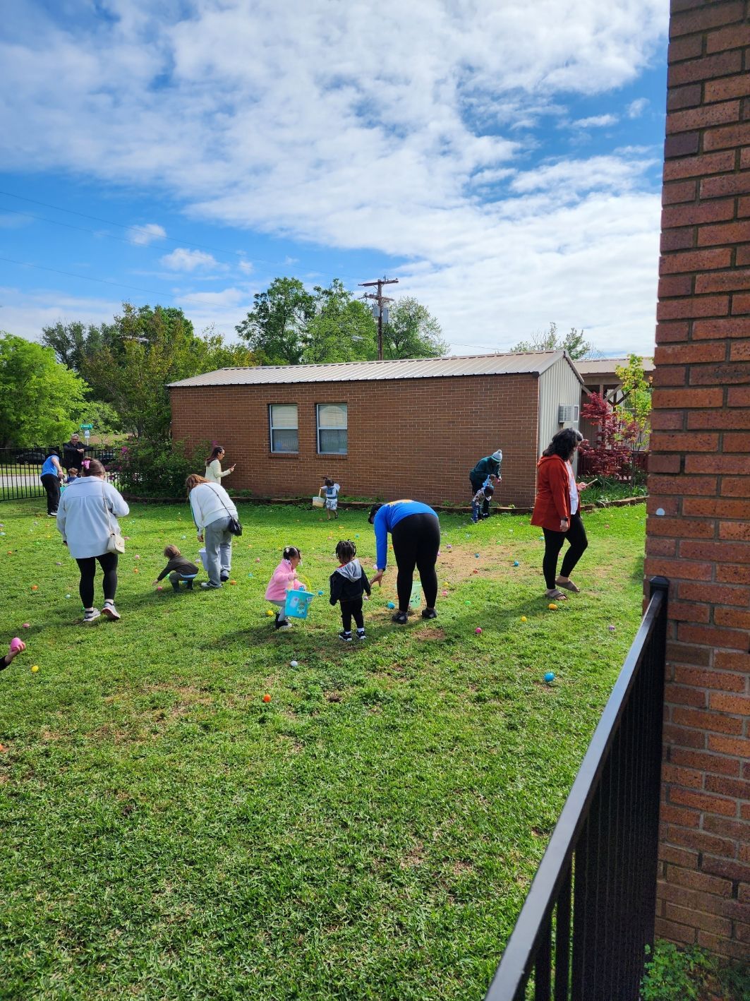 People and children search for eggs in a grassy yard under a blue sky during an Easter egg hunt.