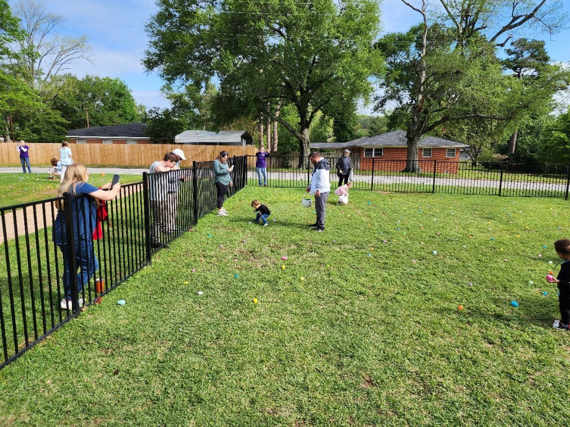 Children search for eggs in a grassy yard during an Easter egg hunt, with adults watching along a black fence.
