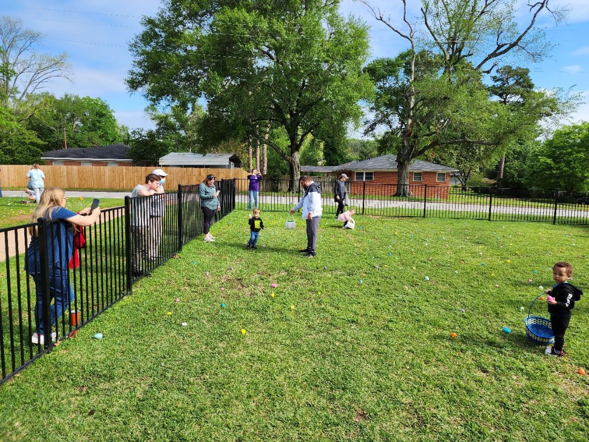 People stand near a black fence surrounding a grassy area with children.
