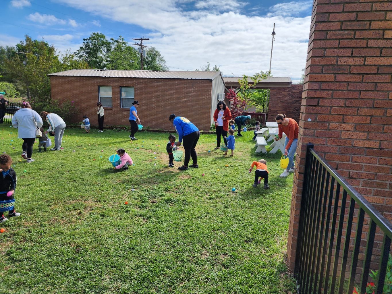 Children and adults search for Easter eggs in a grassy yard. Brown buildings are in the background.