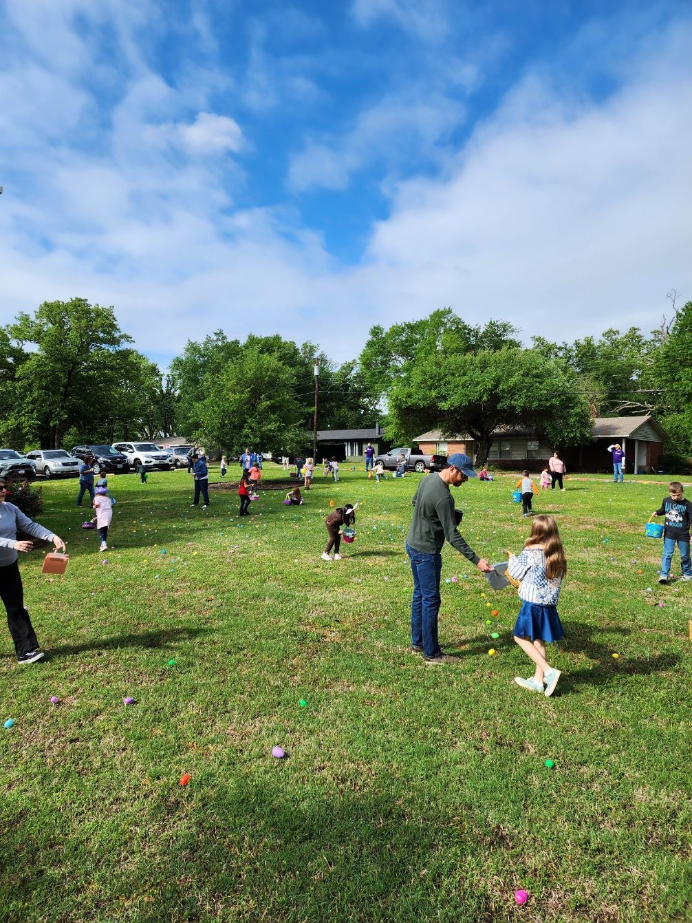 People, including children, hunt for Easter eggs in a grassy field under a cloudy sky.
