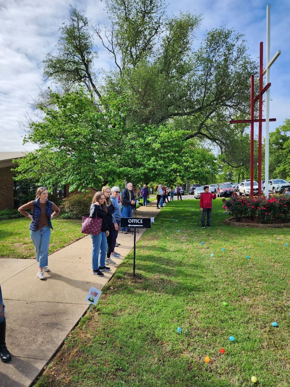 People line up on a sidewalk for an event, near a church with a large cross and a tree, scattered eggs on the grass.