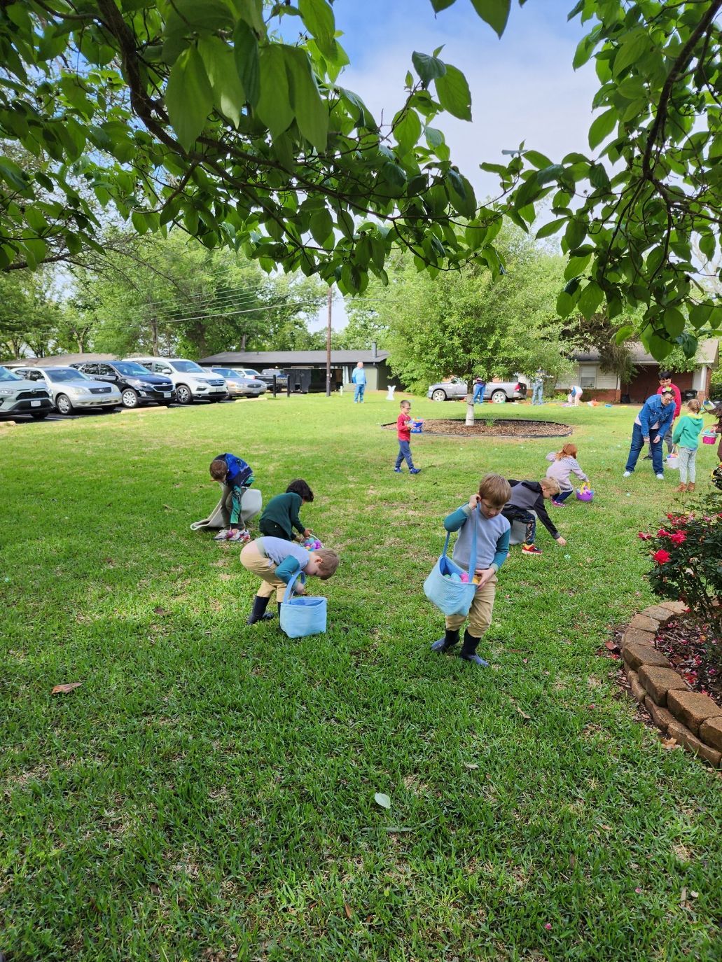 Children hunt for eggs in a grassy yard, with blue baskets. Other people stand nearby.