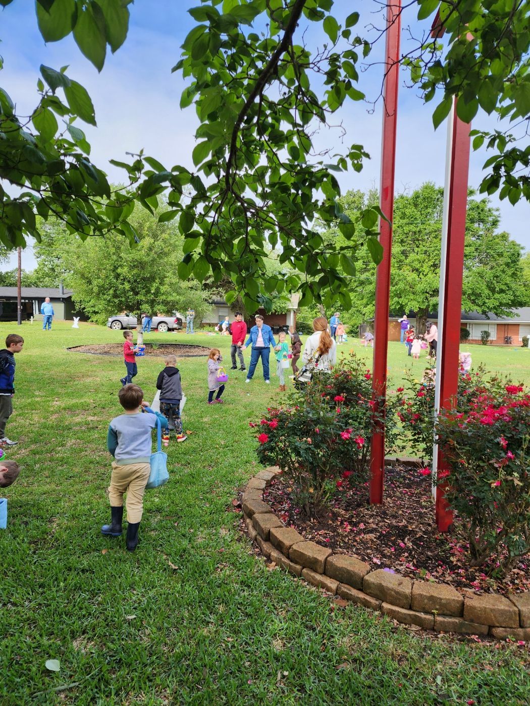 Children hunt for Easter eggs in a grassy yard, under a partly cloudy sky. Red poles and rose bushes in the foreground.