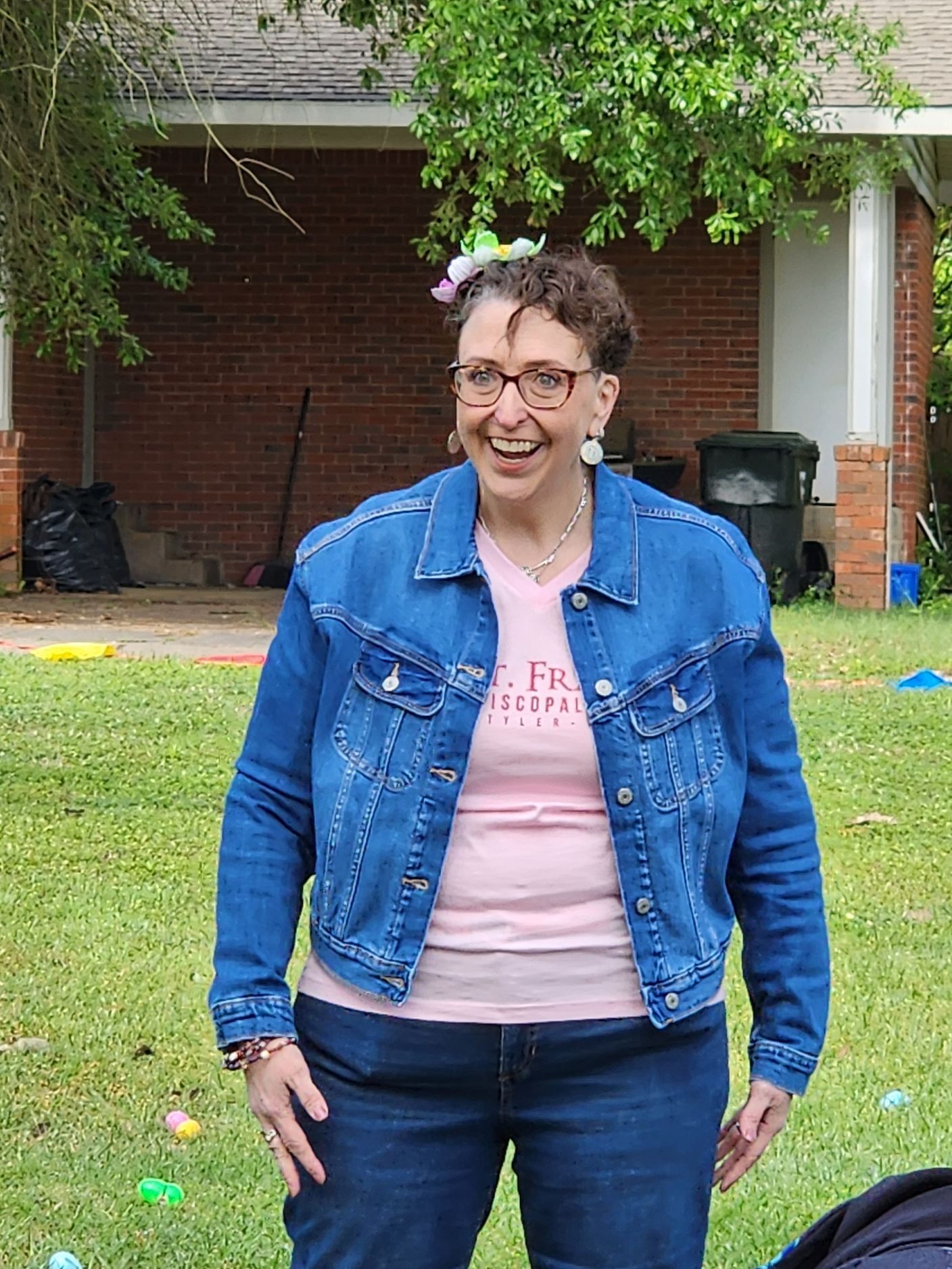 Woman in denim jacket, smiling, wearing glasses. Outdoors, green grass, house in background.