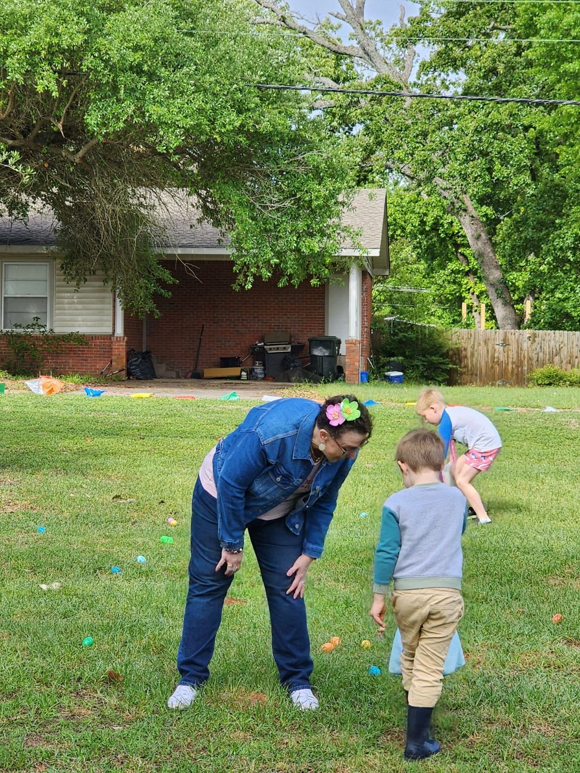 Woman and two children search for Easter eggs in a grassy yard, house in background.