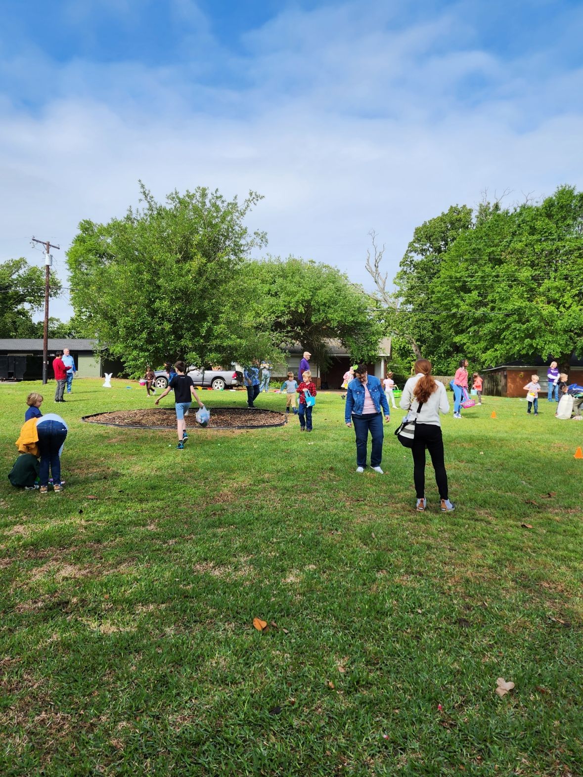 People searching for eggs in a grassy yard under a partly cloudy sky.