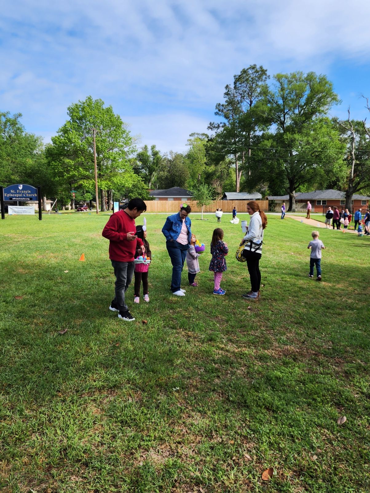 People and children gather on a grassy field, possibly at an outdoor event. Trees and buildings are in the background.