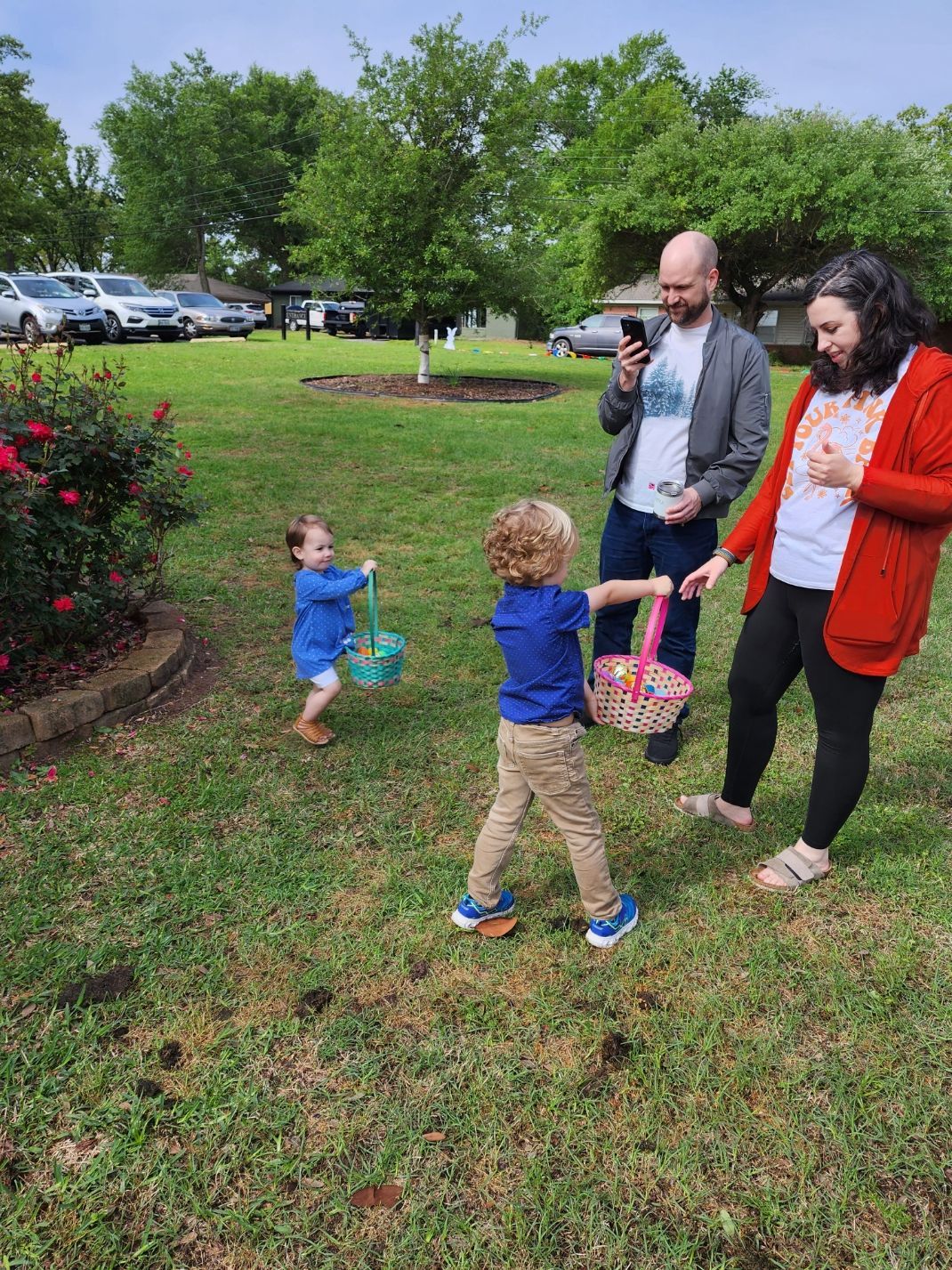 Two children with baskets, a woman, and a man in a grassy yard, likely an Easter egg hunt.