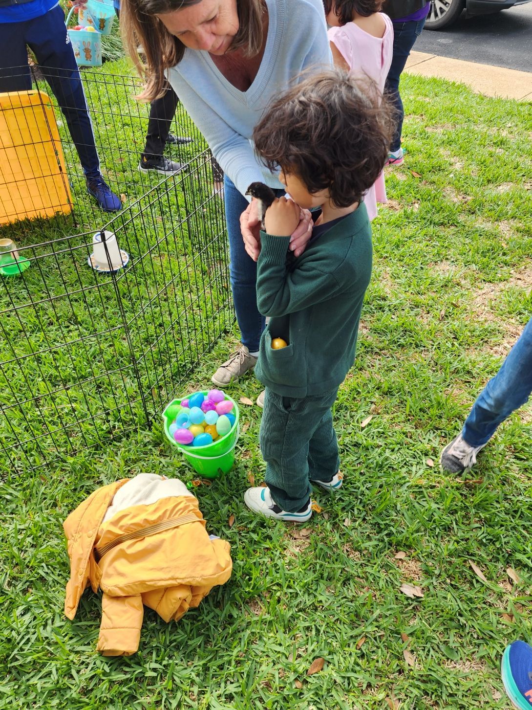 Child and adult at Easter egg hunt: child looking at hand, woman nearby, green bucket of eggs, yellow jacket on grass.