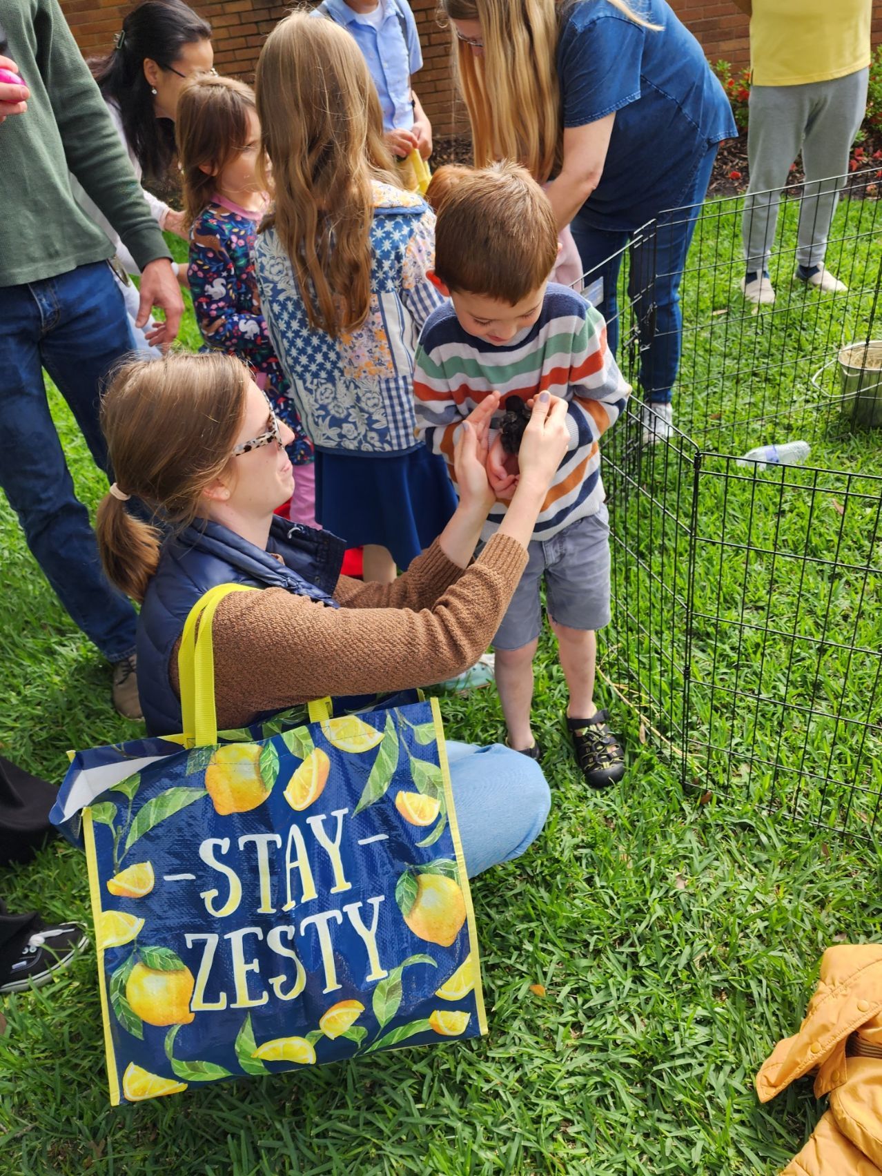 Woman helping child hold a small animal outside, surrounded by others on a grassy area.