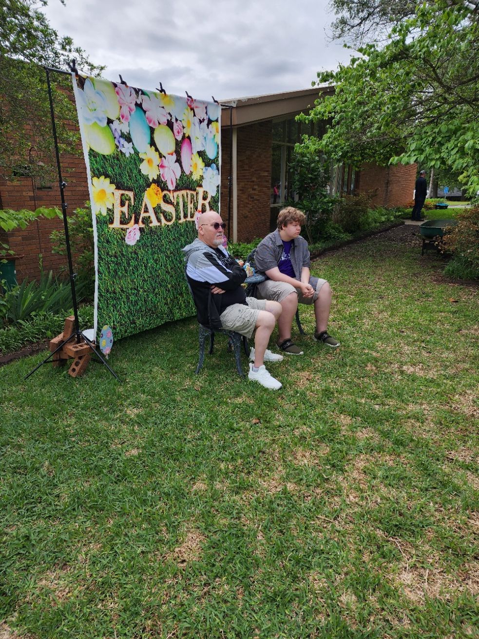 Two people sit on chairs in front of an Easter-themed backdrop in a yard.