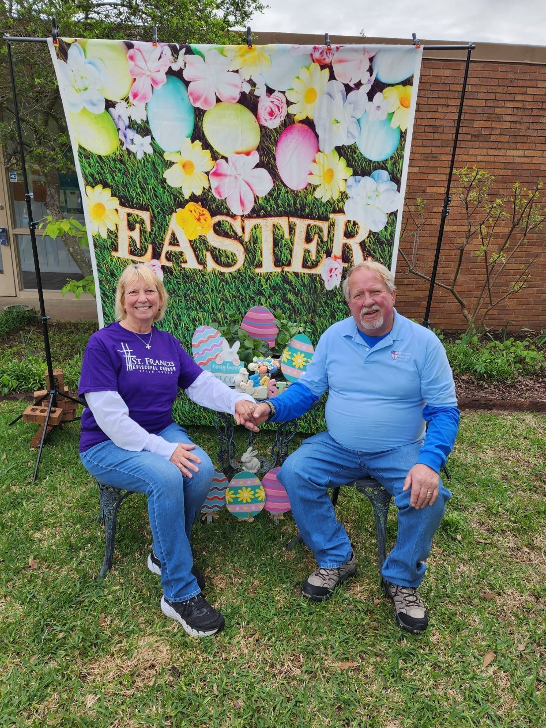 Two people holding hands in front of an Easter-themed backdrop. They're seated on chairs outside.