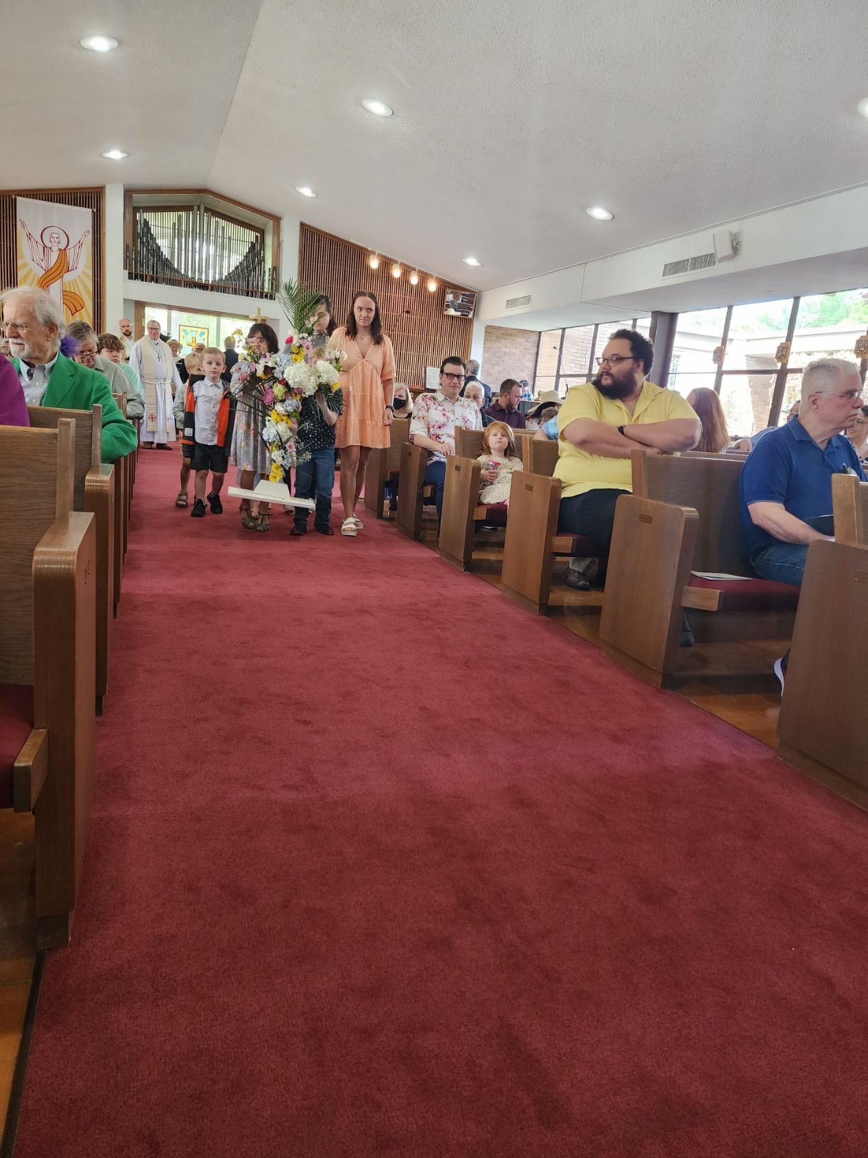 People in church, some with flowers, walk down the red carpeted aisle. Wooden pews line sides.