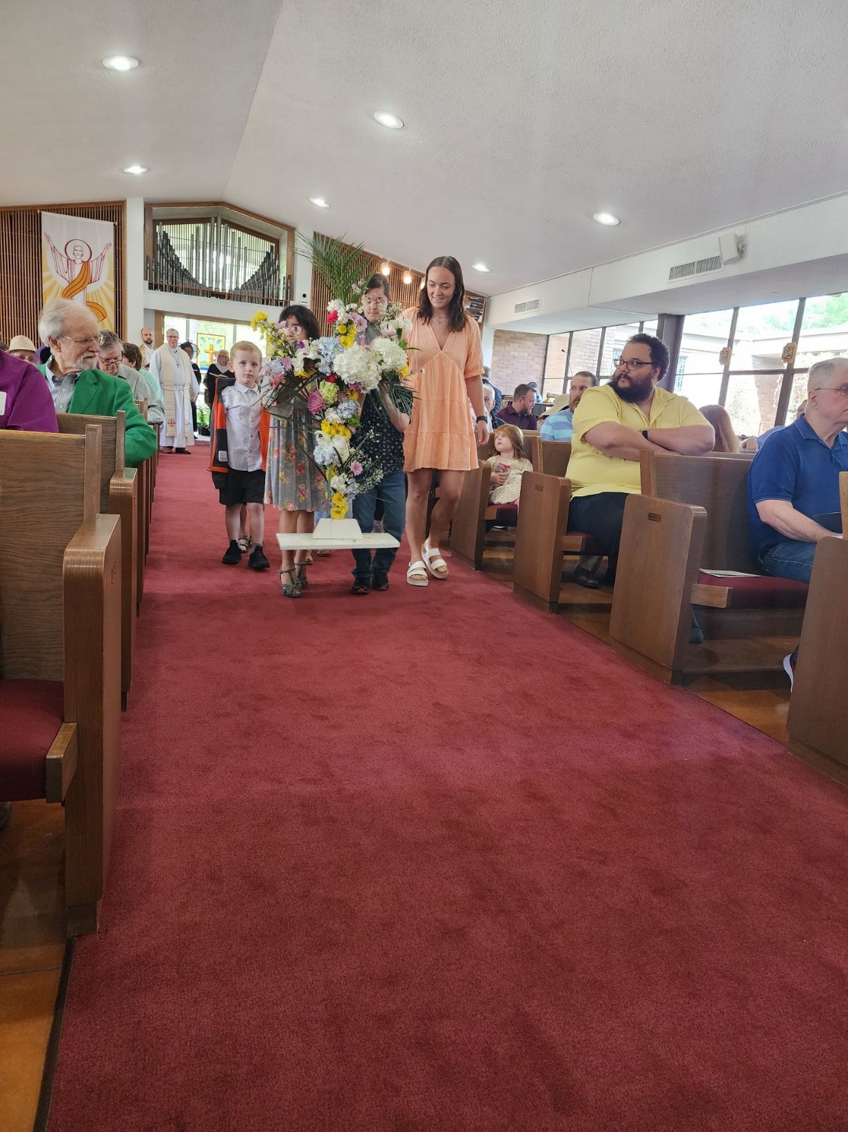 Woman carrying flowers walks down aisle of church, followed by children; people seated in pews.