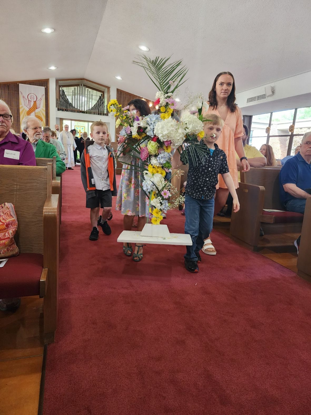 Two boys and a woman carry a flower-covered cross down a church aisle.