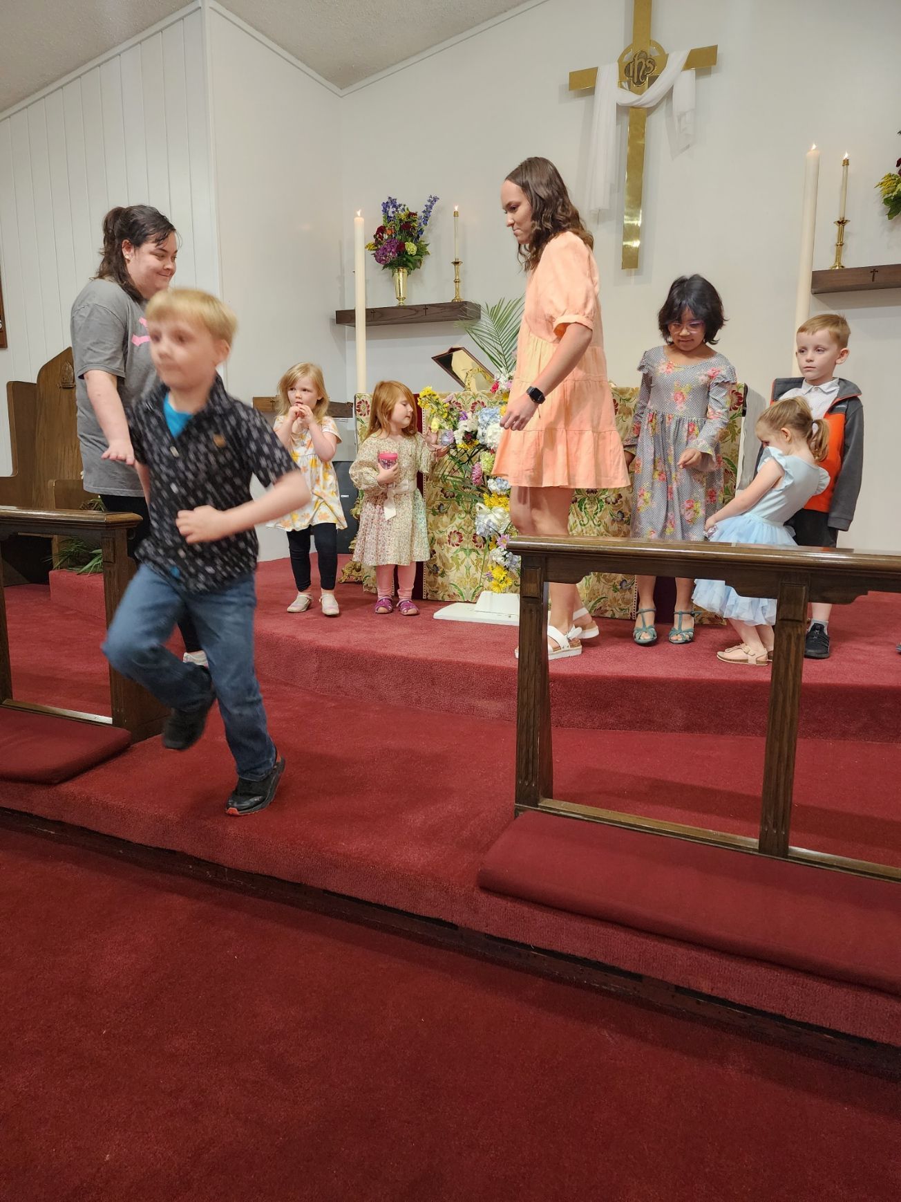 Boy running down church aisle; children and adults on stage; red carpet.