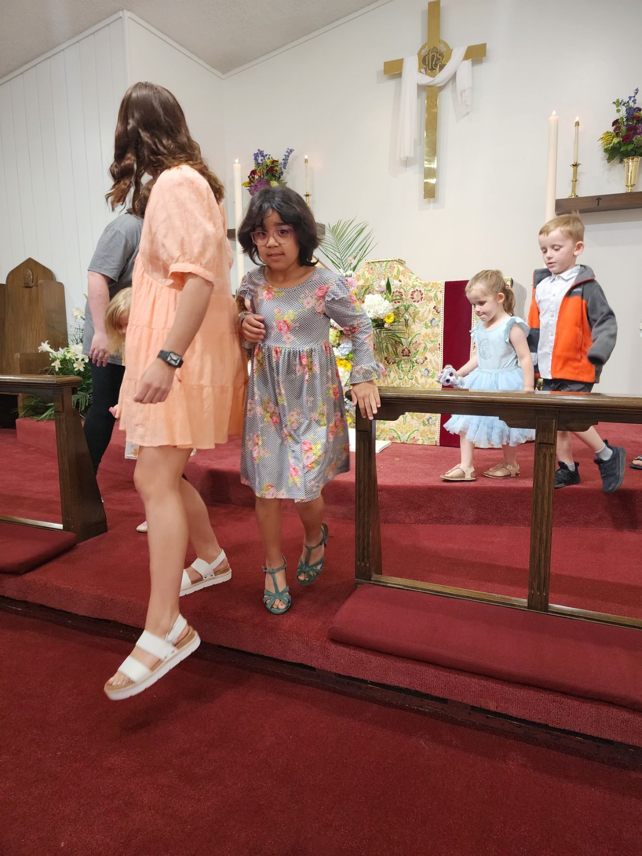 Children walking down a church aisle on a red carpet. A cross and candles are in the background.