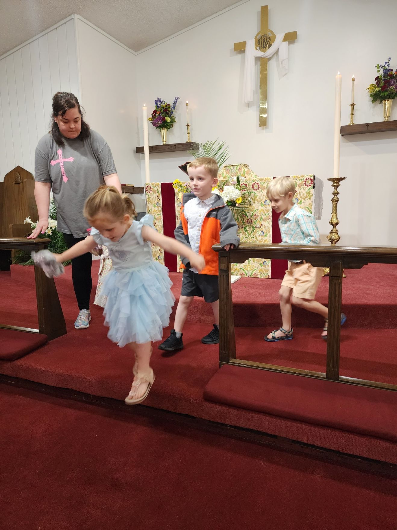 Woman helps three children step over a barrier in a church. Red carpet, floral backdrop, candles.