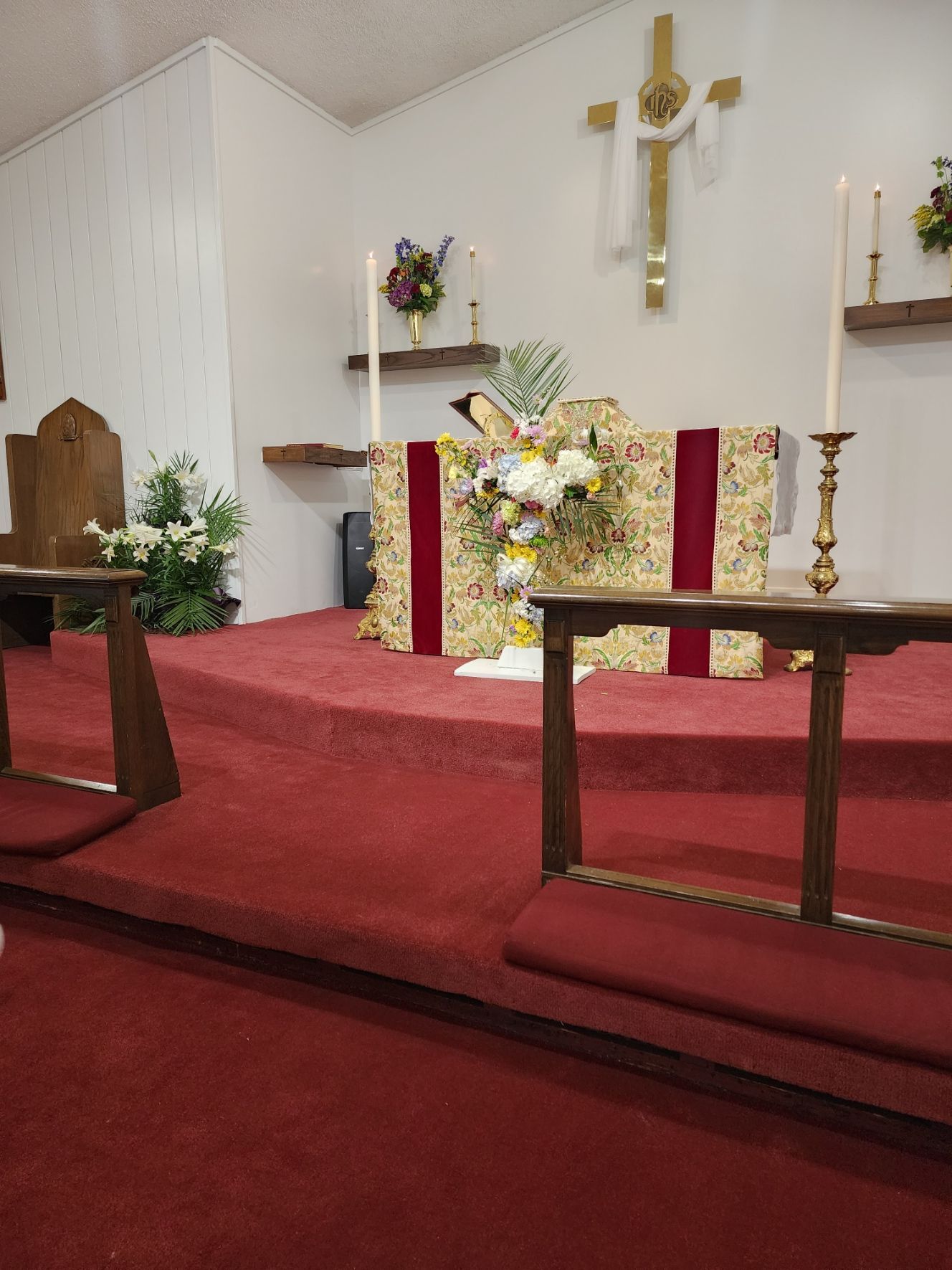 Church altar with flowers, candles, and a gold cross on a red carpeted platform.