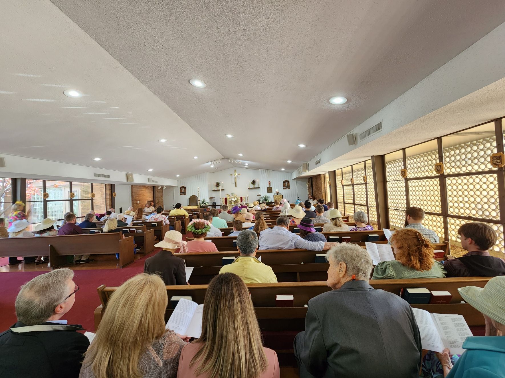 People seated in pews during a church service; natural light streams through side windows, with a cross visible in the center.