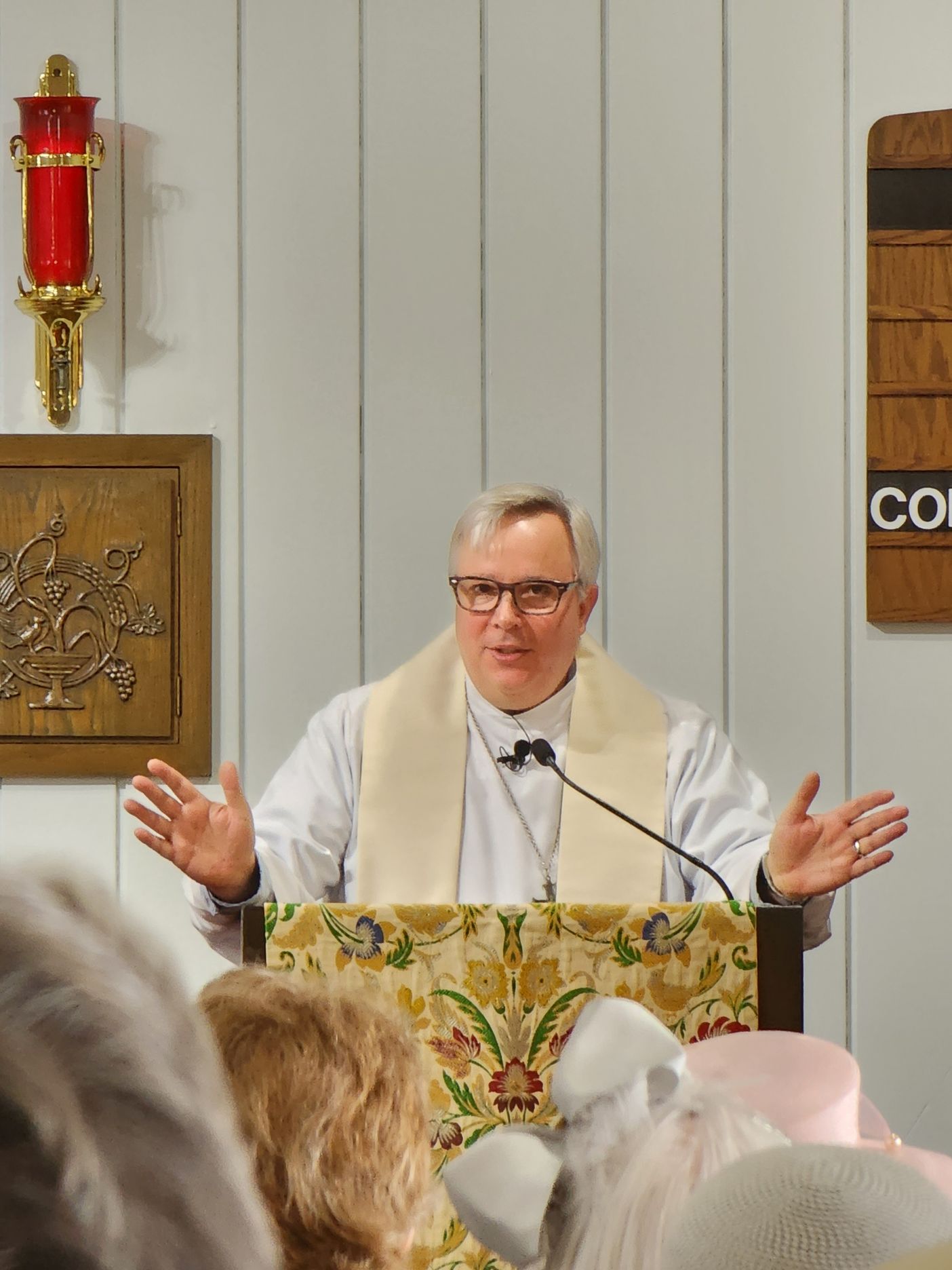 Clergyman speaking from a pulpit, arms outstretched. White collar, glasses, indoors.
