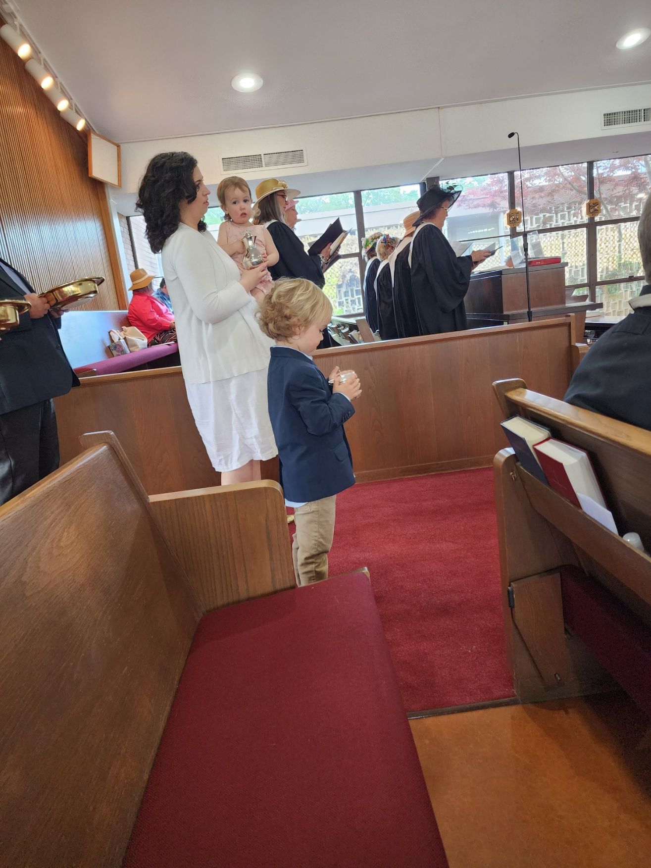 A church service with a boy in a blazer standing, mother holding a child, and clergy at the altar.