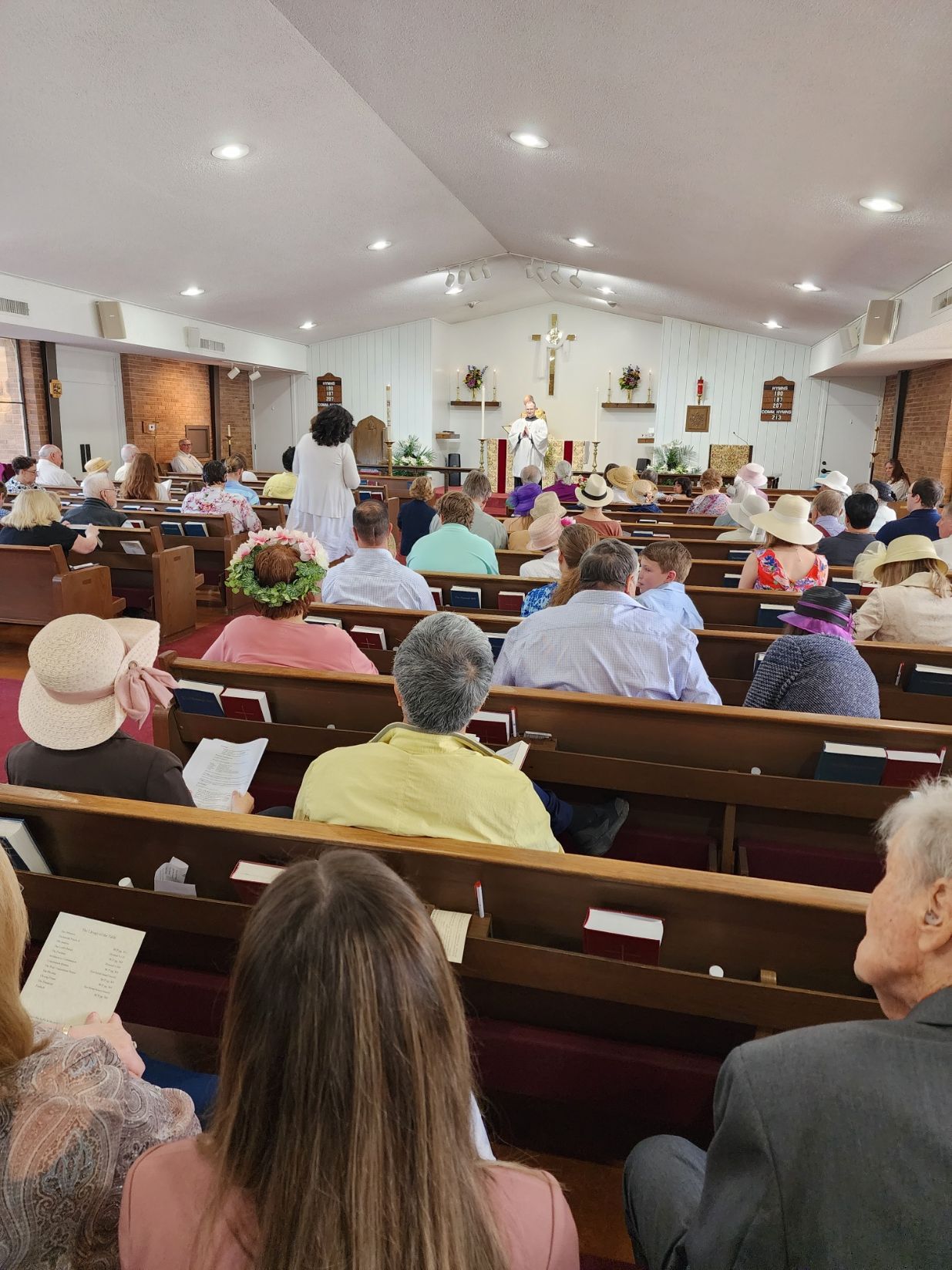 People seated in church pews, facing altar. Several wear hats. Sunlight streams through windows.