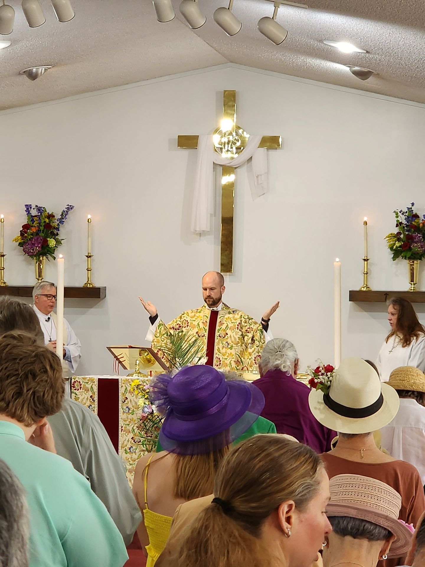 Clergy member leading service; people in the congregation. Altar with gold cross, candles, and flowers.
