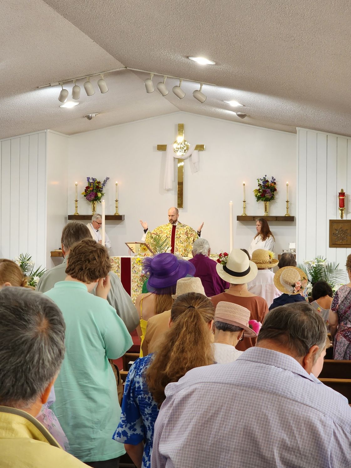 Church service with a priest at the altar, attendees facing forward. Sunlight, cross, candles.