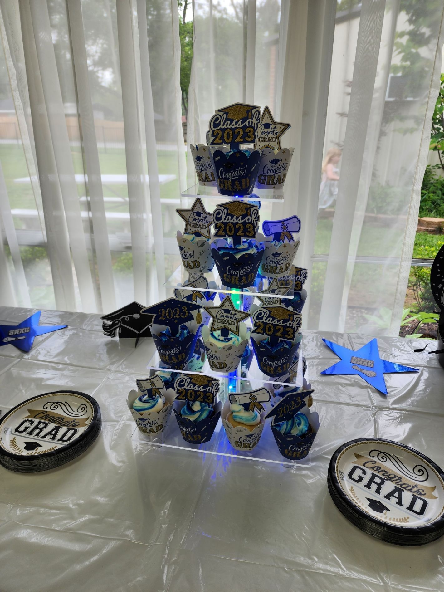 Graduation cupcakes on a tiered stand, blue and white decor with star accents, two plates, outdoors.