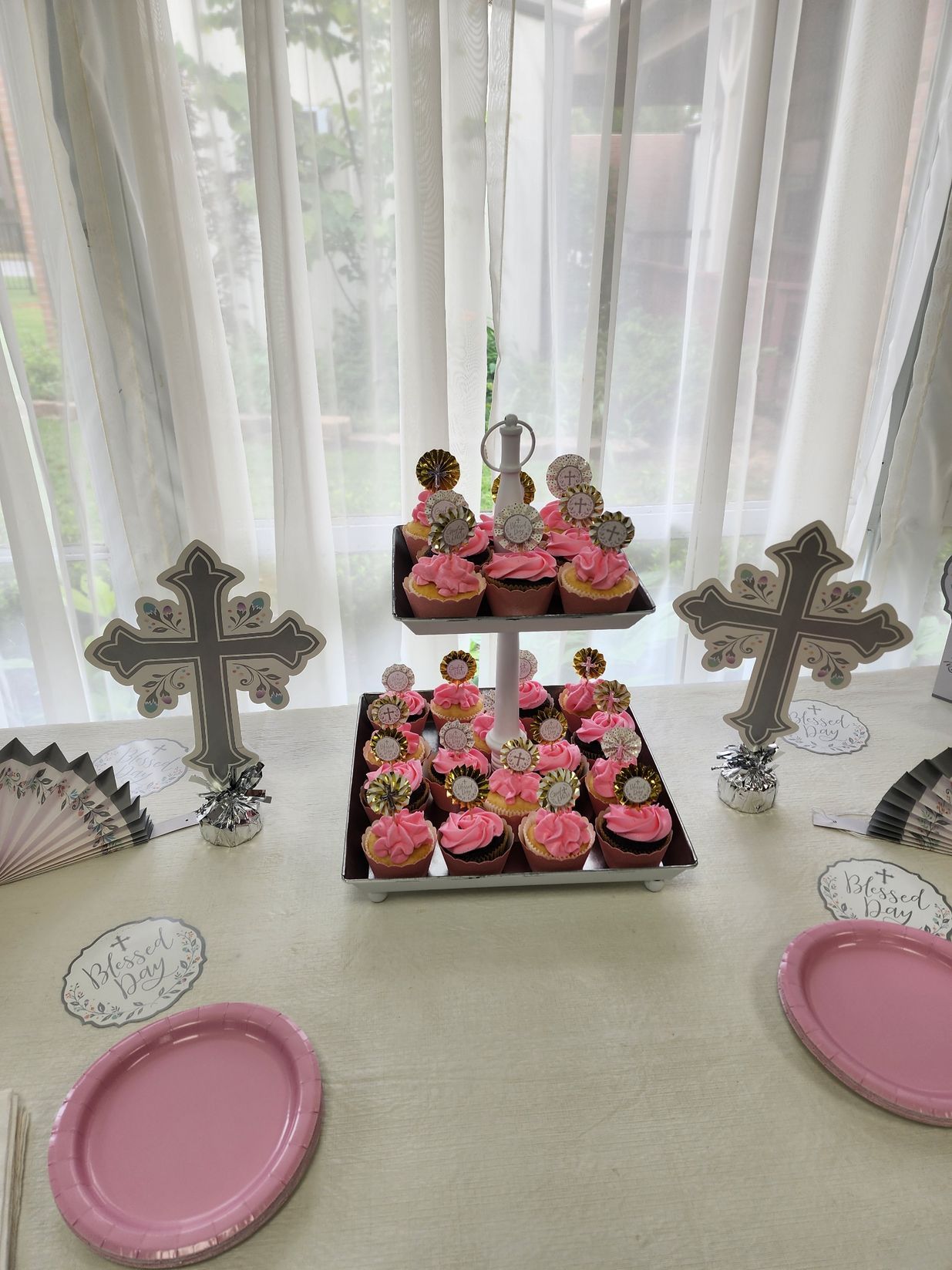 Cupcakes with pink frosting on a tiered tray, decorated for an event, on a table. Two decorative crosses.