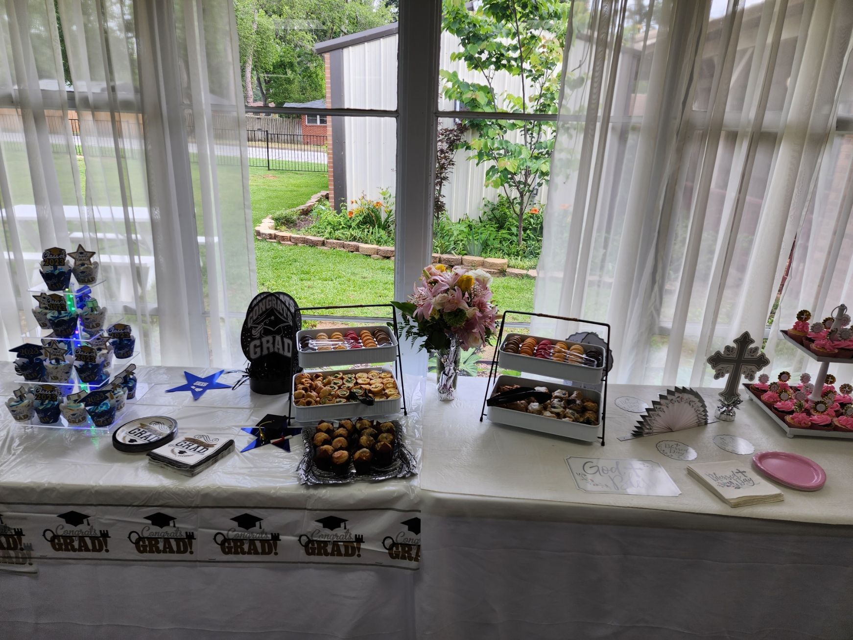 Graduation party dessert table with cupcakes, treats, and floral arrangement near a window.