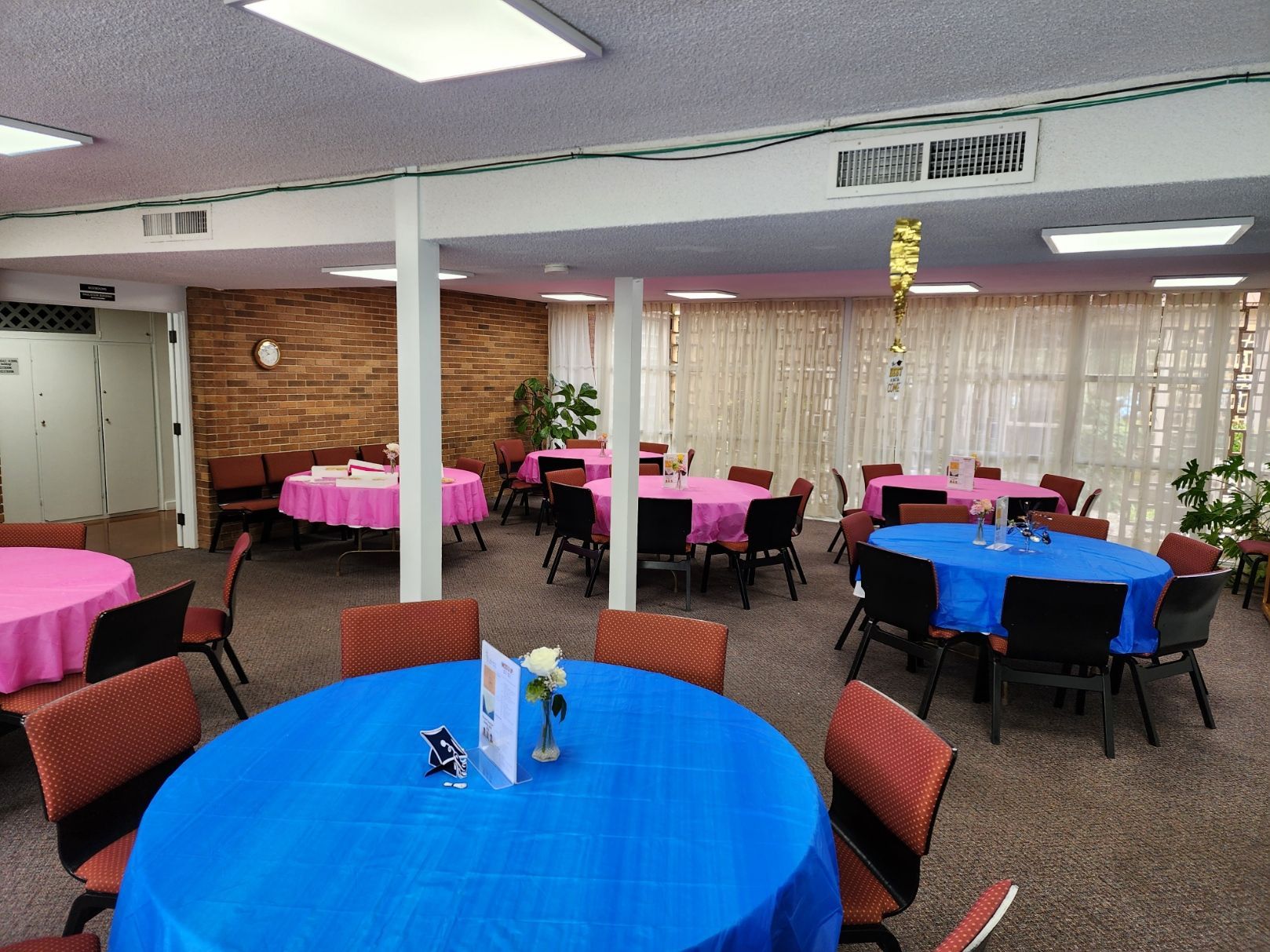 Round tables covered in pink and blue tablecloths in a room, set for a party.