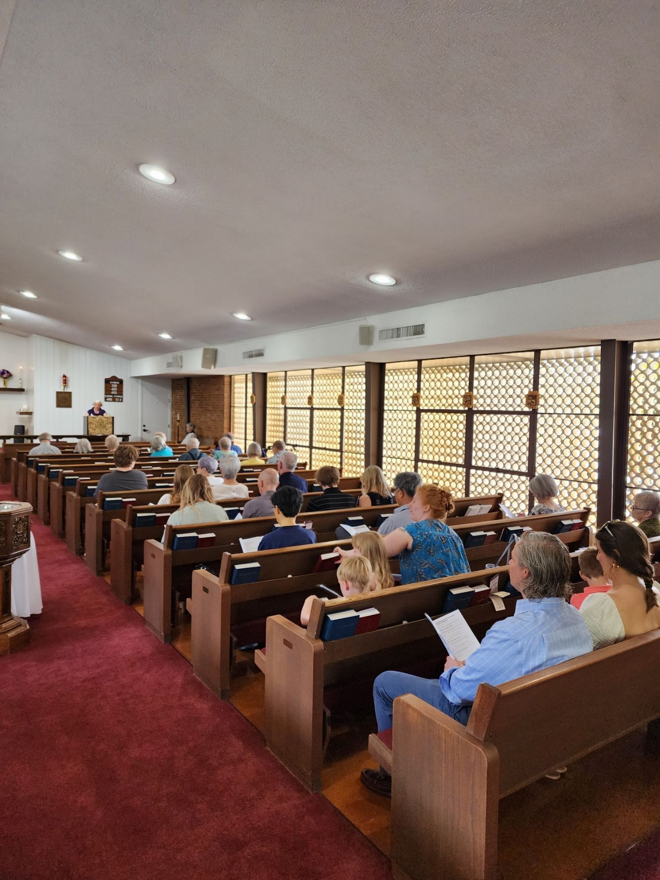People seated in pews in a church. Brown pews, red carpet, and brown patterned windows are visible.