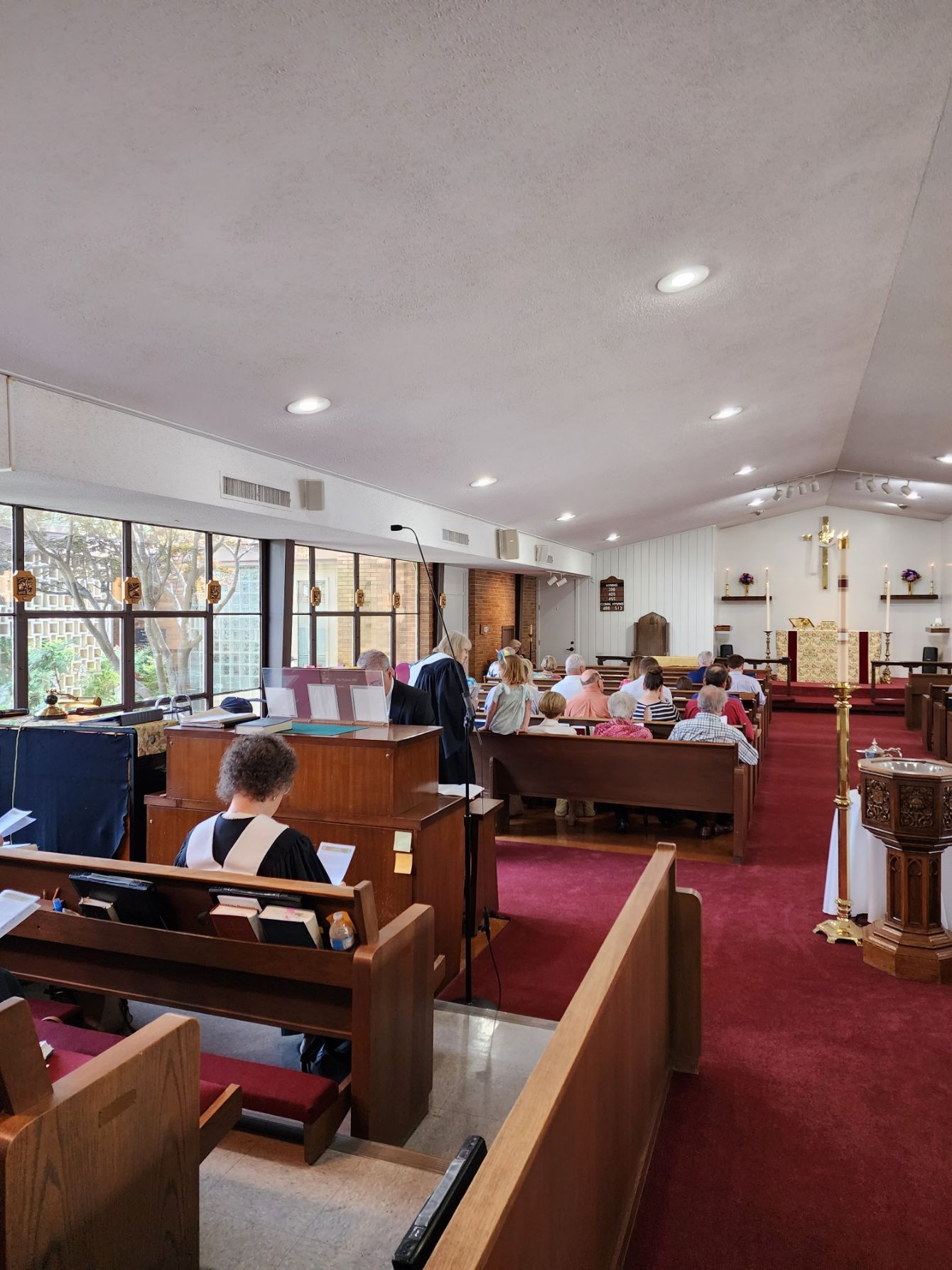 Interior church view: People seated in pews, facing a front altar with a cross. Red carpet, light walls.