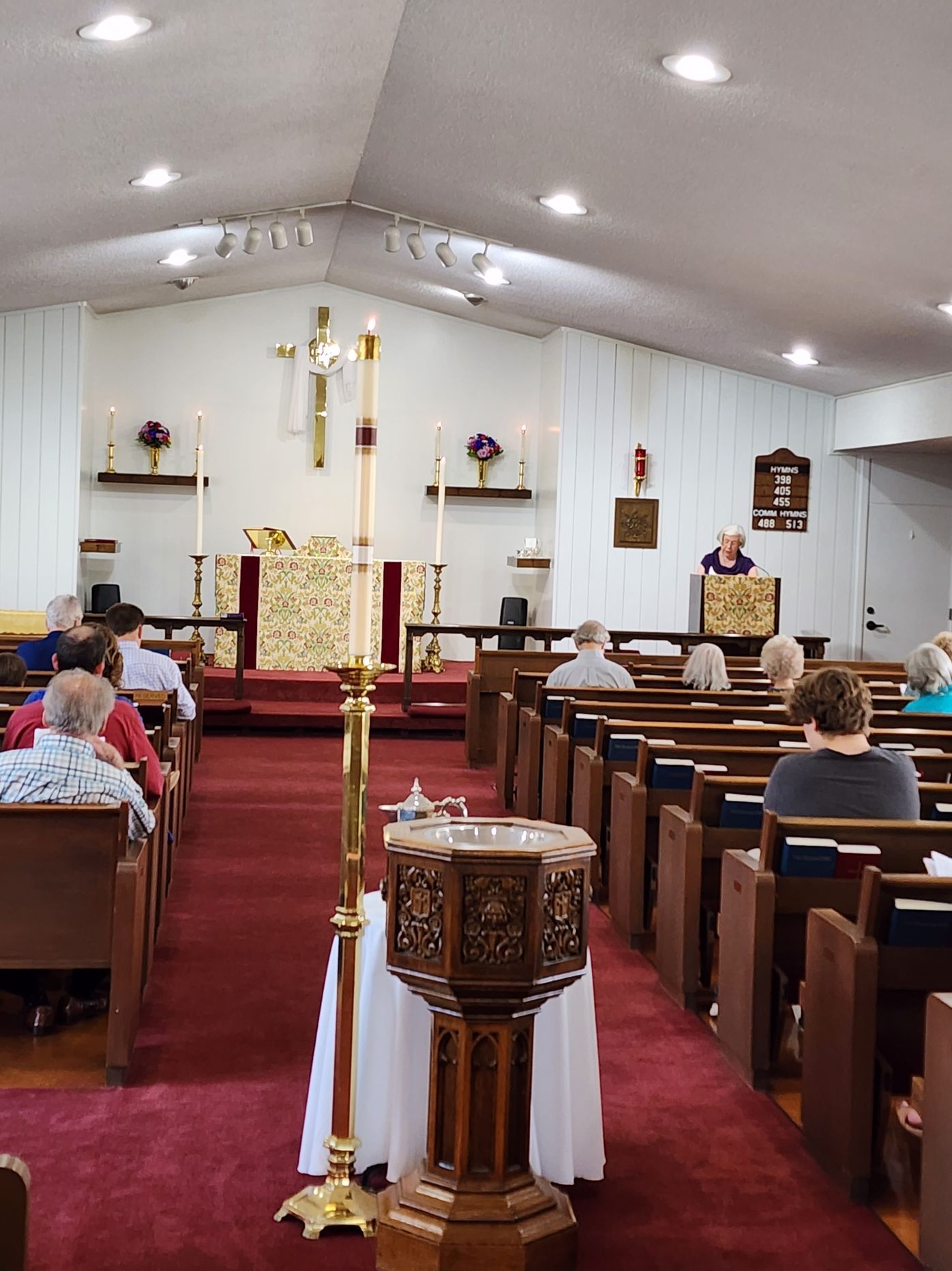 Interior of a church during a service. People sit in pews facing a pulpit. Lit candles and a cross are visible.