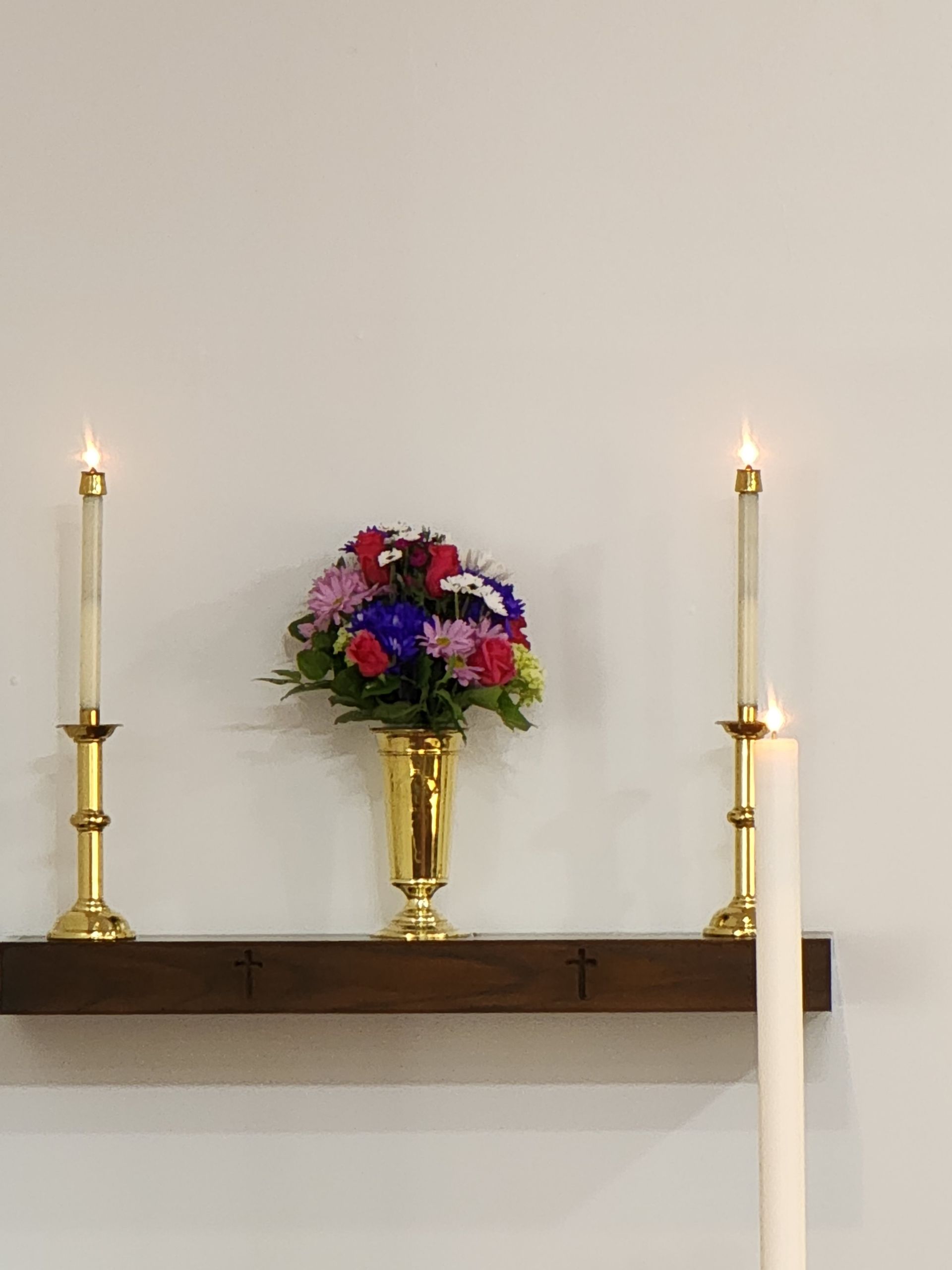 Flowers in gold vase between lit candles on a wooden shelf against a white wall.