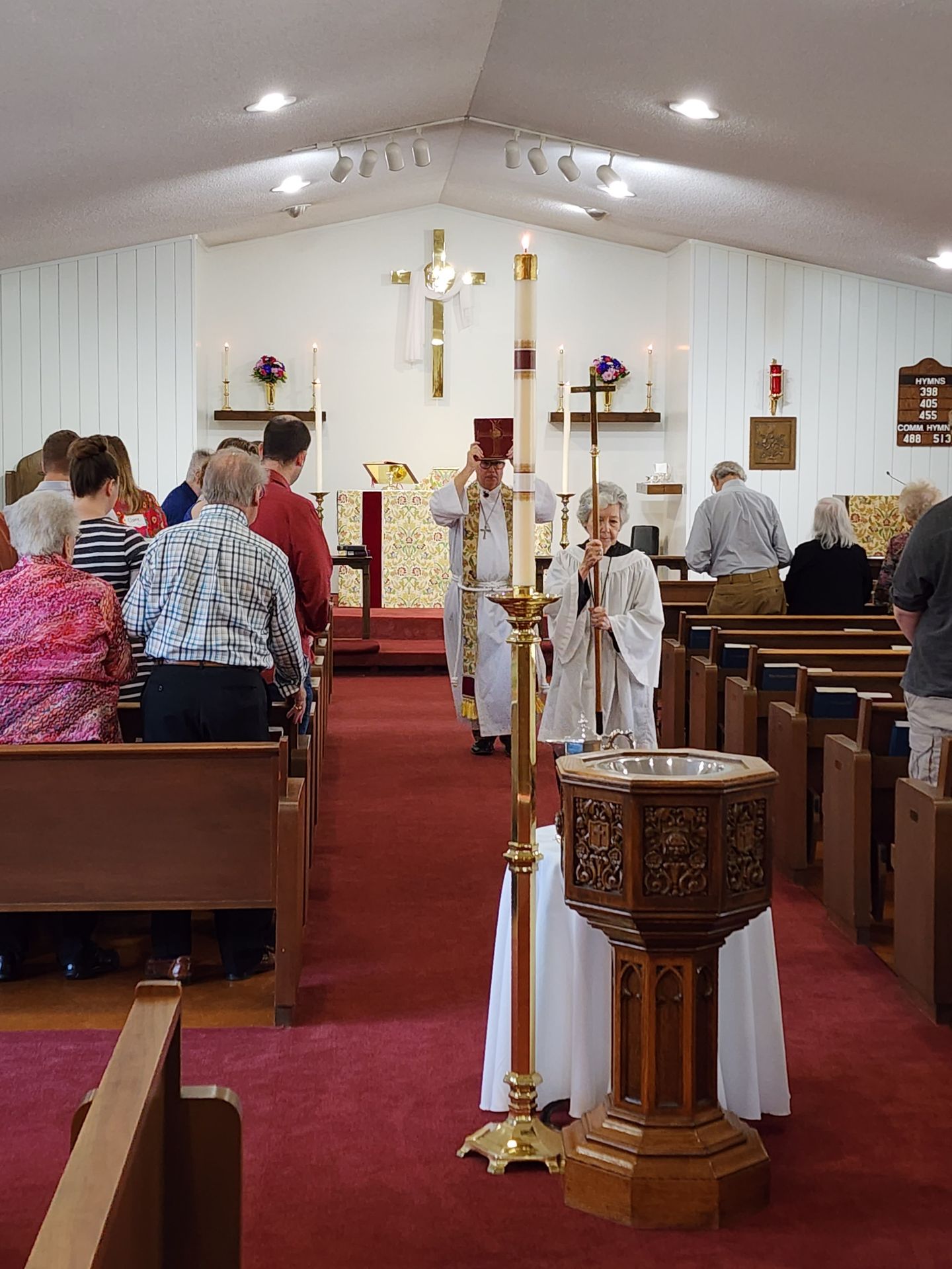 People in church, priest leading procession, lit candles, red carpet, wooden pews.