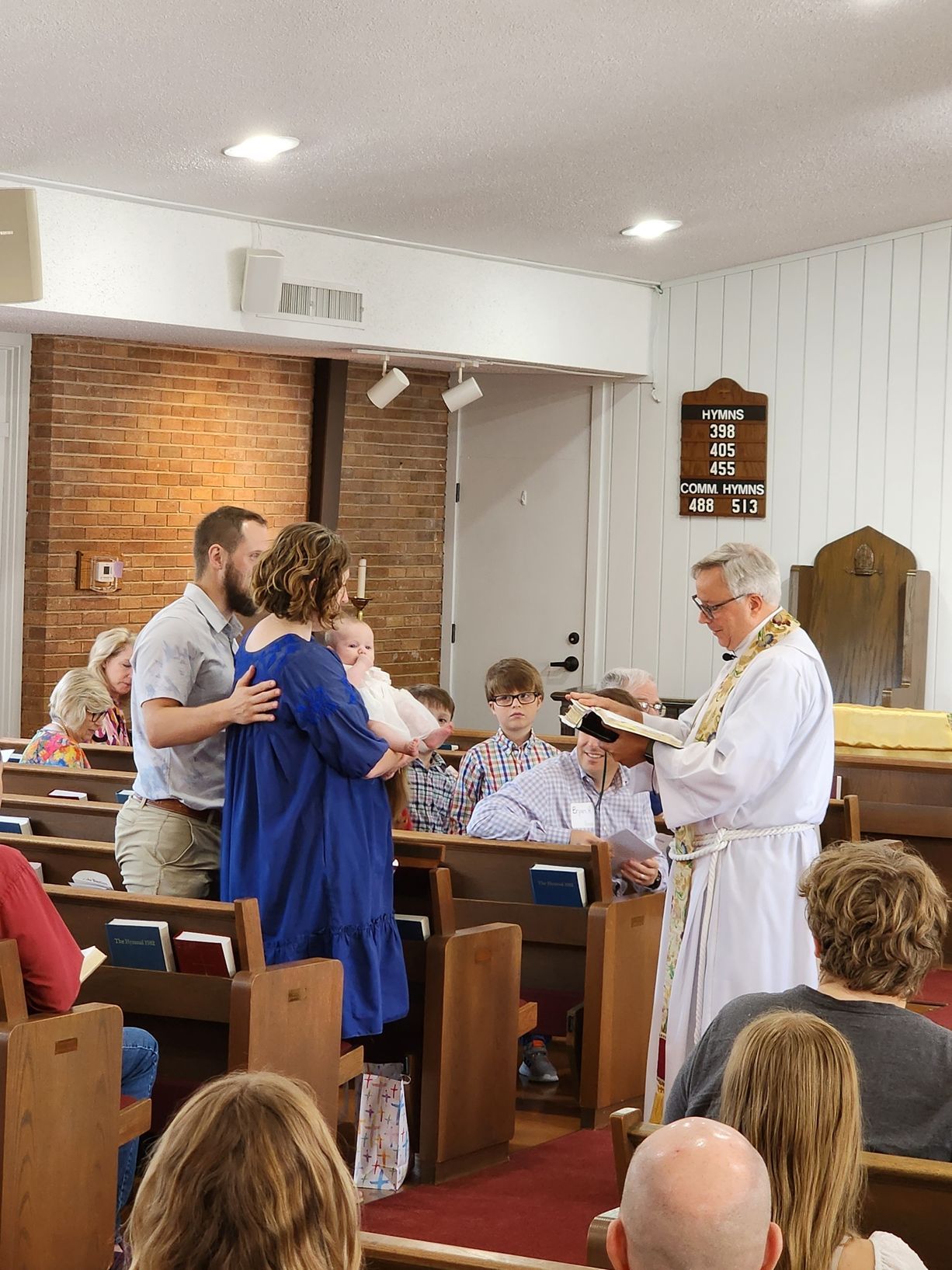 People at church ceremony, priest with Bible, family in wooden pews.