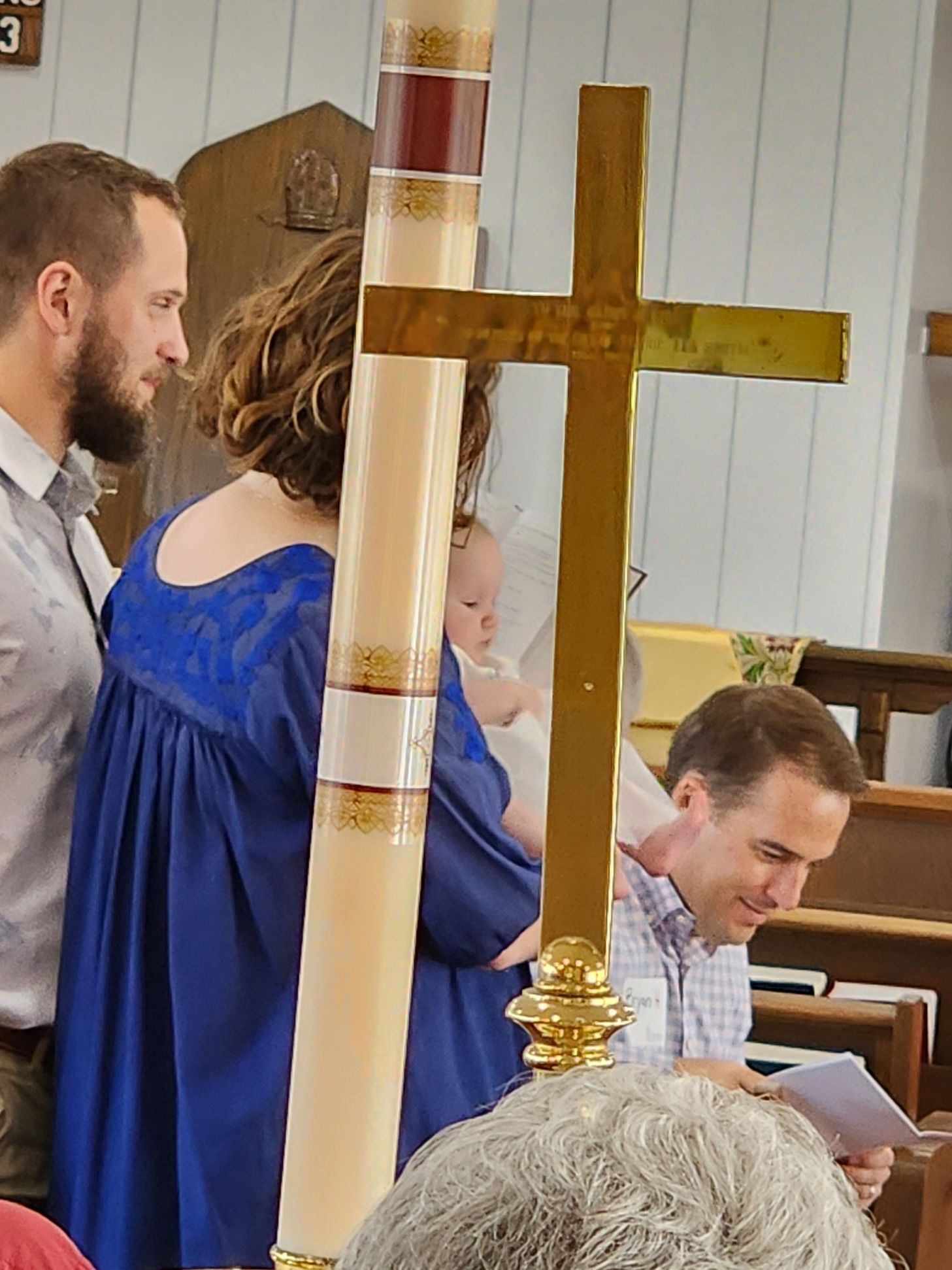 Baptism ceremony: people near candle and cross, woman holds baby, man looks at papers.