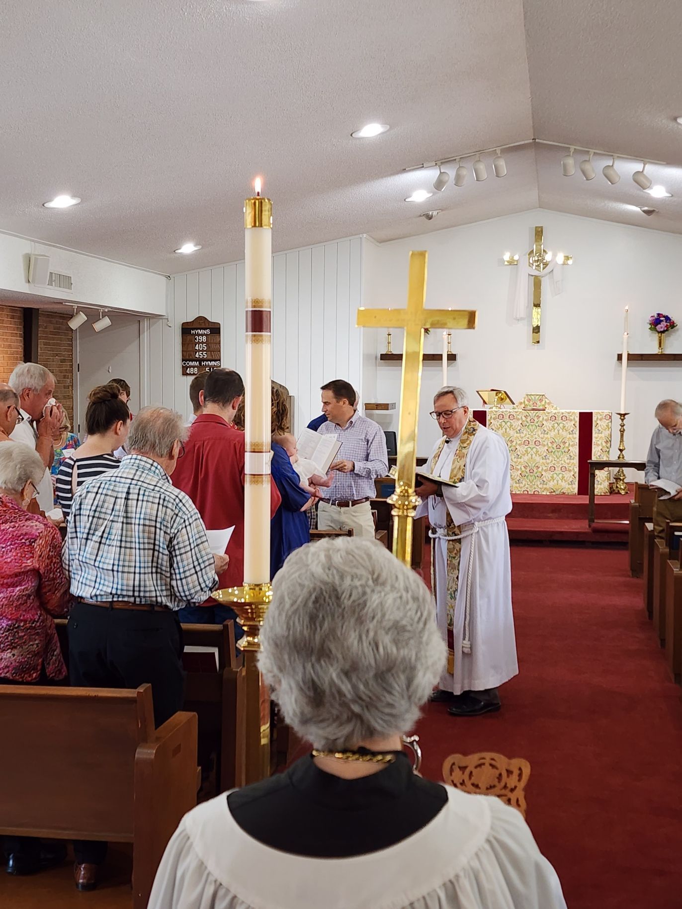 Church service: Priest in white robes, golden cross, tall candle. Congregation singing, red carpet.