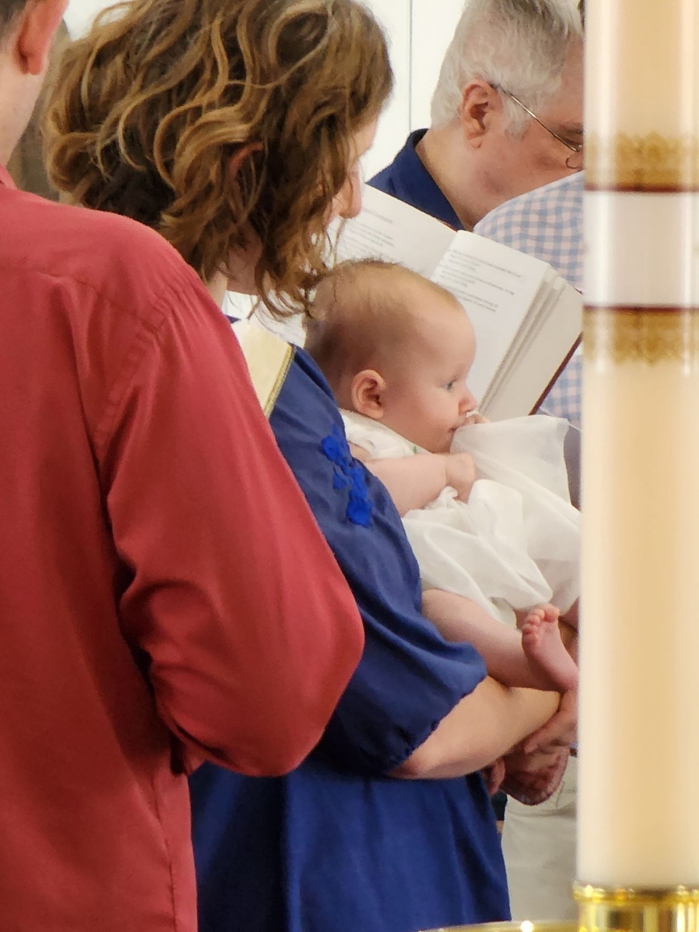 Woman holding a baby in a white dress, at a religious ceremony. Others are nearby.