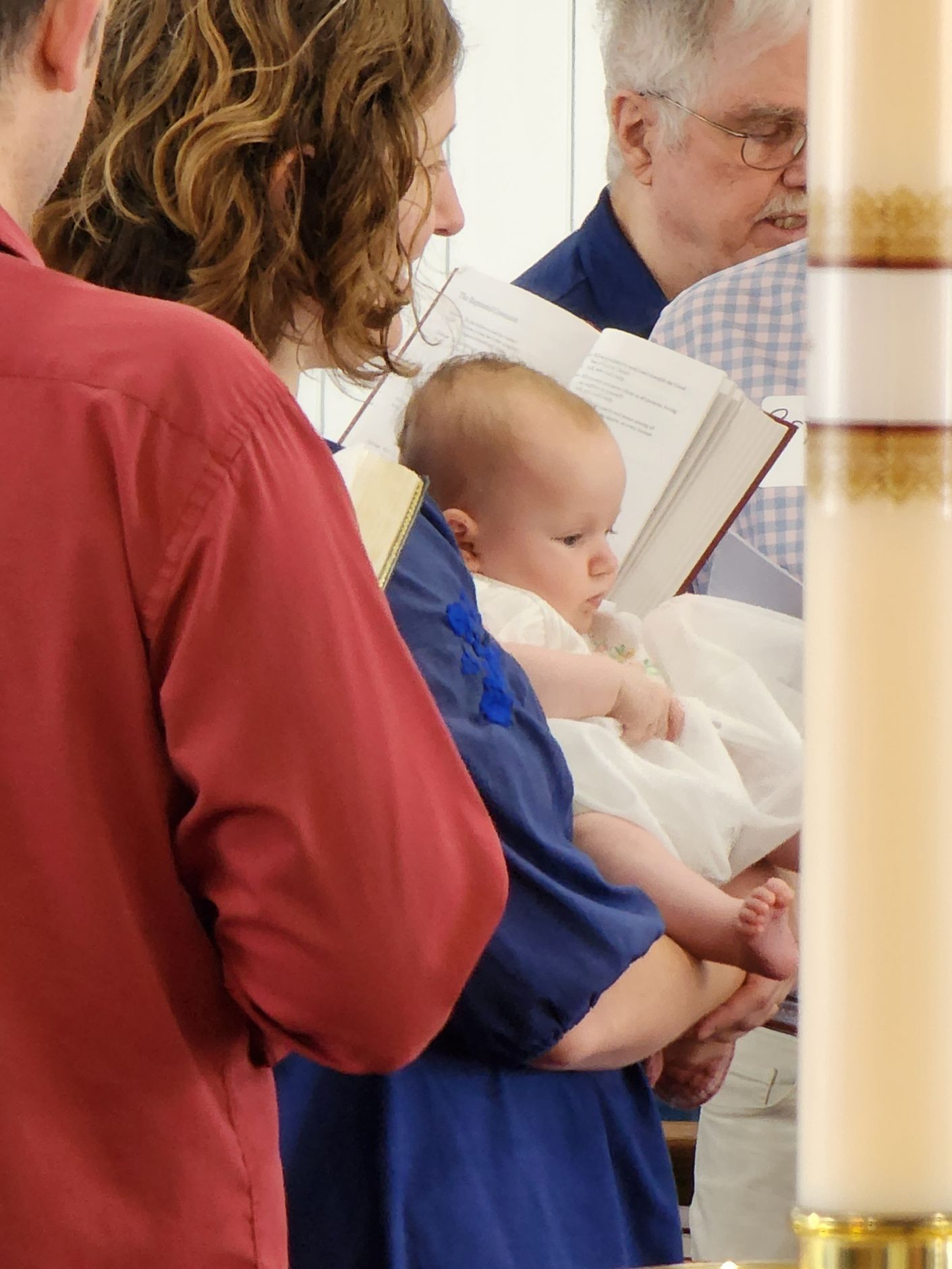 Baby in white dress held by person in blue, at ceremony with others.