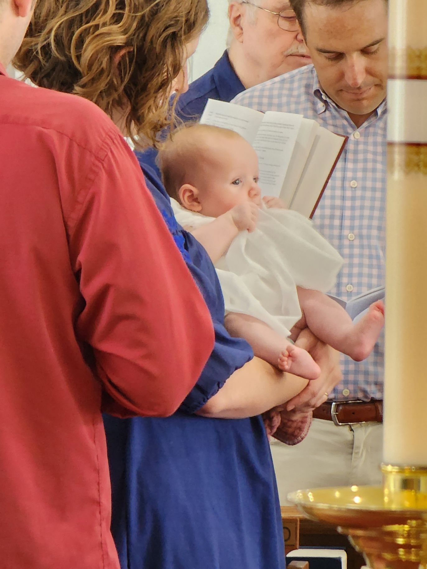 A baby in white held by a person in blue during a baptism ceremony, with other people nearby.