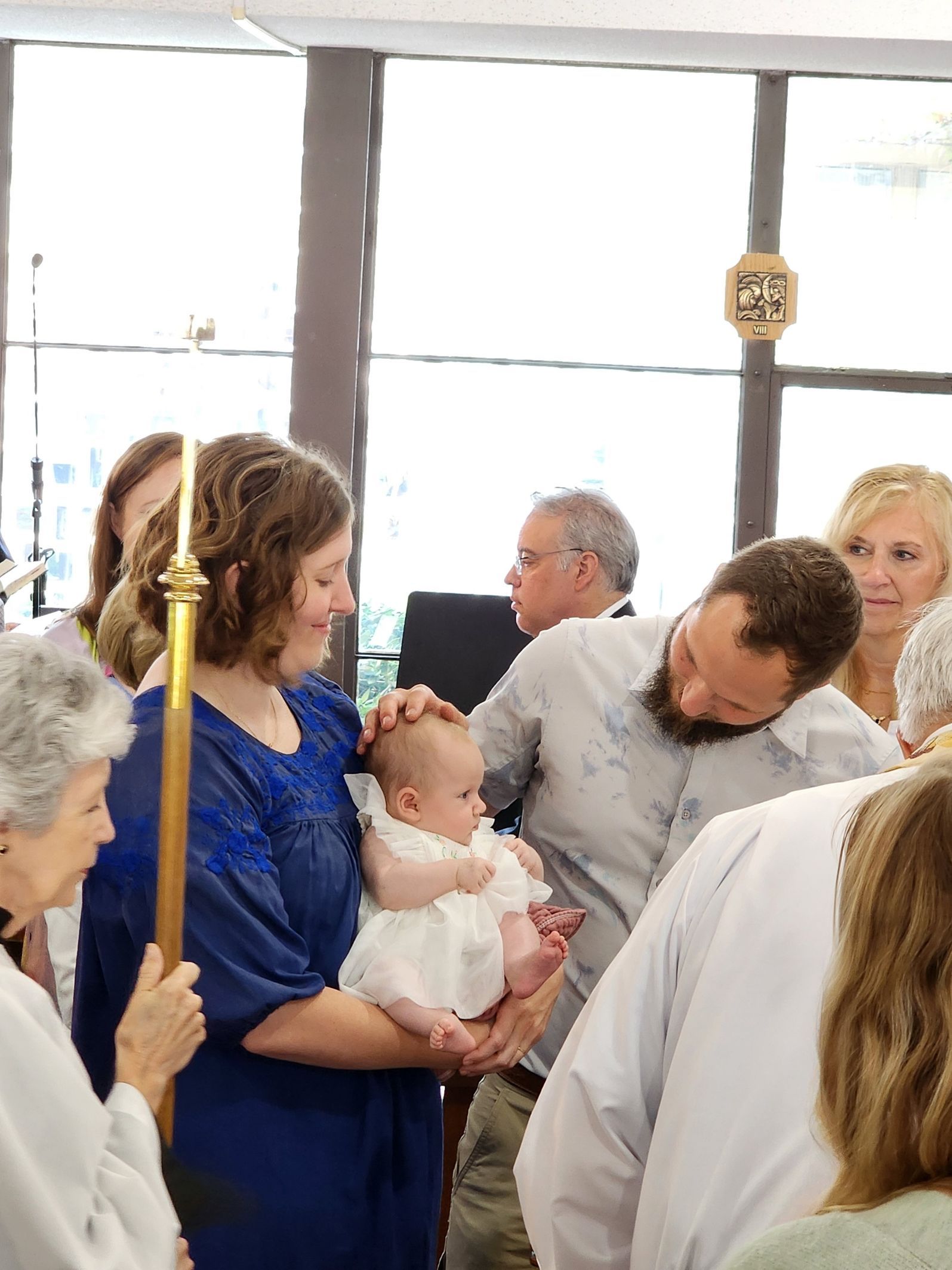 Baby held for baptism ceremony, with family and clergy present. Sunlight streams in through windows.