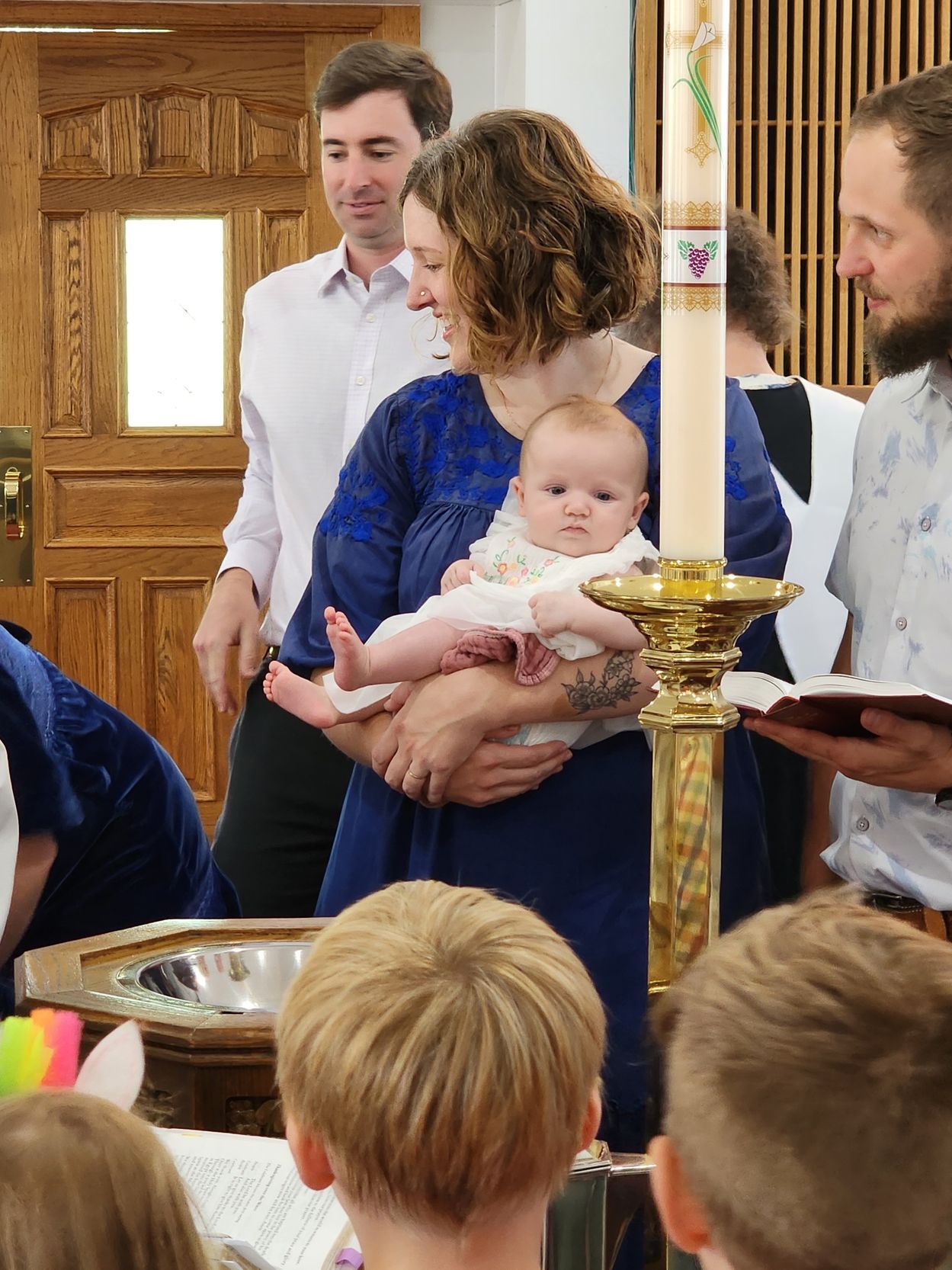 Woman holding baby at baptism; church interior. Other adults nearby.